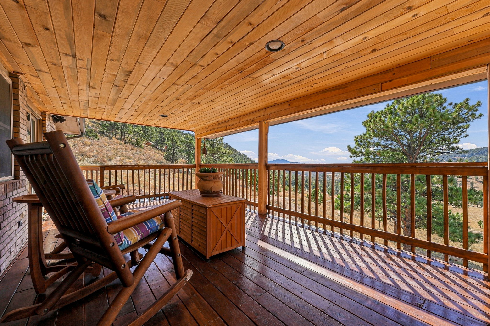 This covered deck offers a serene outdoor living space with a rustic wooden rocking chair and a matching storage chest. The space features a warm wood-paneled ceiling and a wooden railing that frames expansive, panoramic views of the surrounding mountainous landscape and pine trees. The perspective is from the deck looking outward, capturing the interplay of sunlight and shadow across the wooden floorboards.