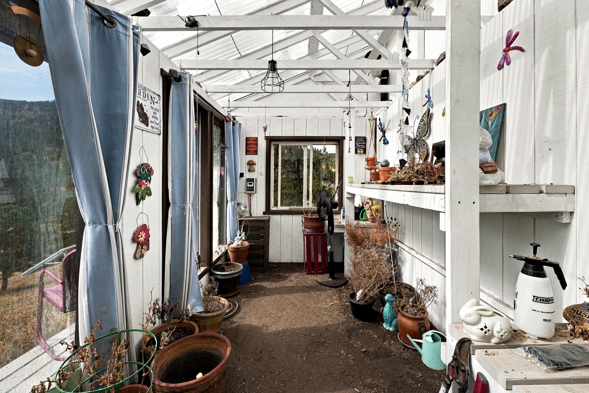 This rustic sunroom or greenhouse space features a dirt floor, white-painted wooden framing, and a translucent roof that allows for ample natural light. The area is decorated with various potted plants, whimsical wall art, and blue-and-white striped curtains, creating a charming, lived-in atmosphere. A window at the far end provides a view of the surrounding landscape, while a workbench and gardening supplies suggest a functional space for plant care.