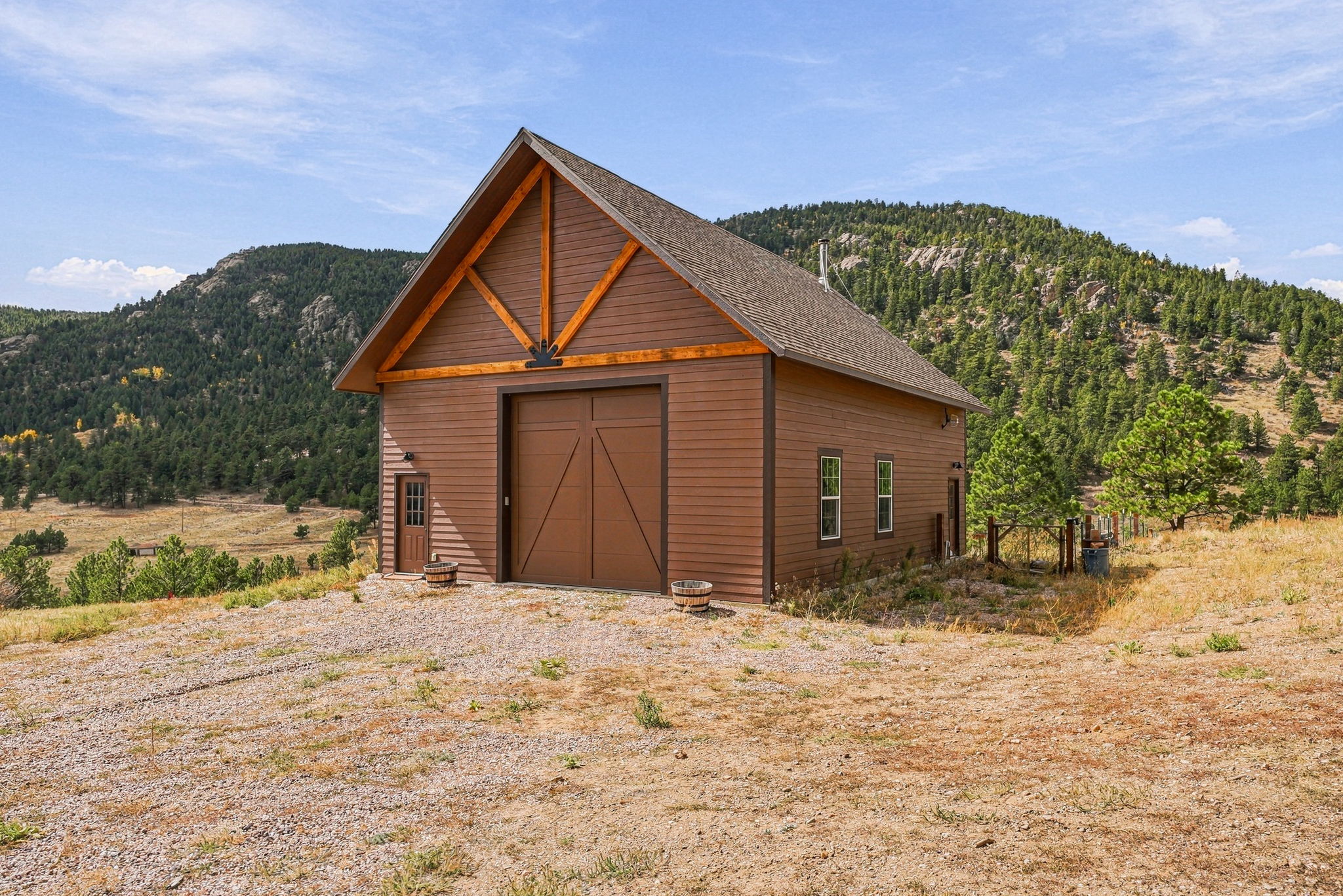 This image showcases a rustic, brown-sided detached garage or workshop set against a scenic mountainous backdrop. The structure features a prominent gable roof with decorative timber trusses, a large central garage door, and a separate side entry door. The surrounding landscape is a dry, gravel-covered lot with native grasses and a dense forest of pine trees climbing the hills behind the building.