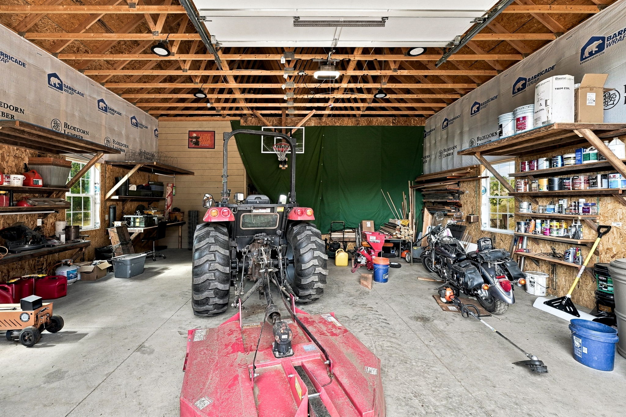 This spacious, unfinished garage features exposed wooden rafters and walls lined with protective wrap, offering a rustic and functional workshop environment. The area is equipped with extensive shelving units filled with various supplies, a tractor with a mower attachment, and a motorcycle parked on the concrete floor. A basketball hoop is mounted on the back wall, which is partially covered by a large green privacy screen, highlighting the versatile utility of this expansive storage space.