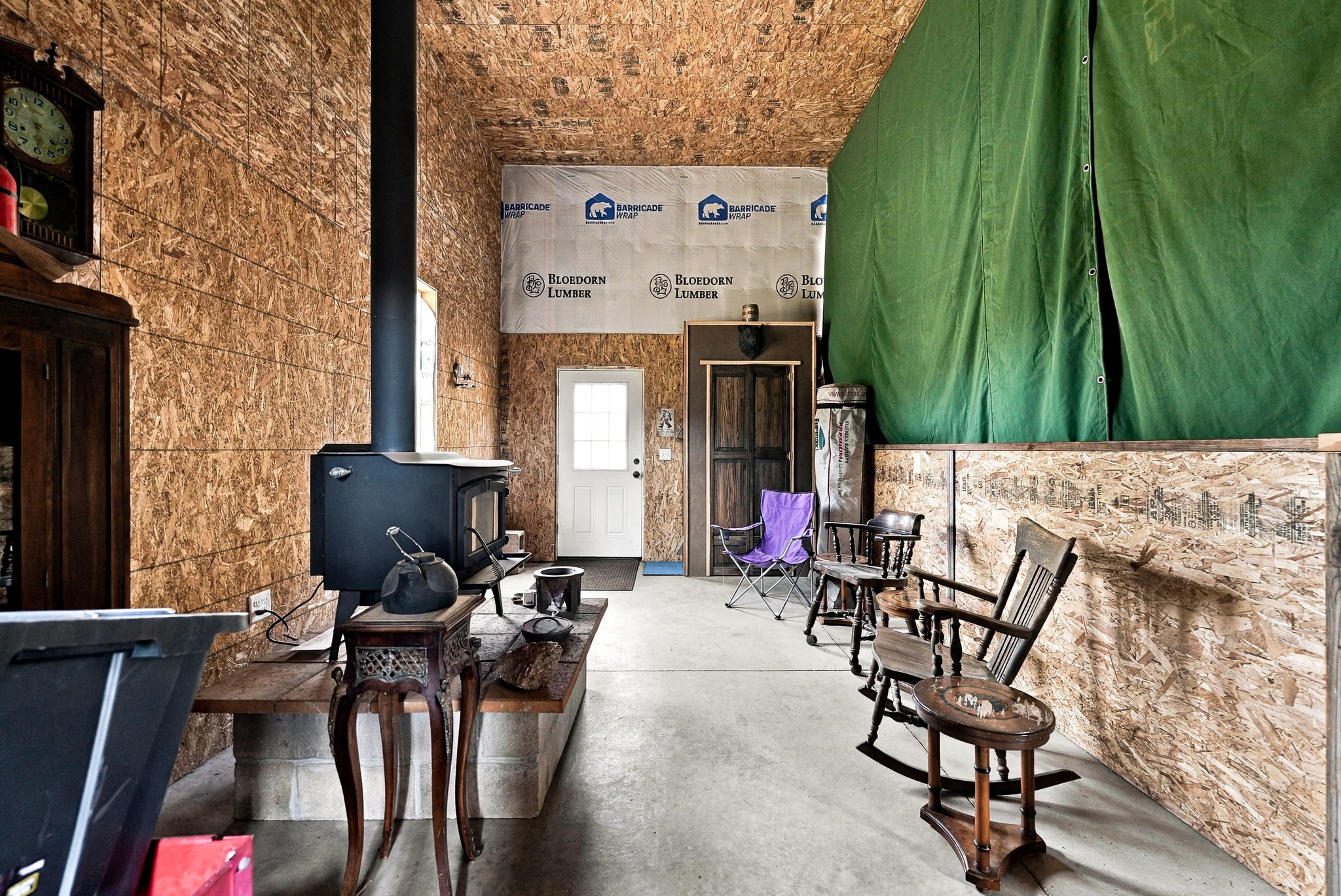 This interior space appears to be a rustic workshop or utility area featuring exposed oriented strand board (OSB) walls and ceiling. A black wood-burning stove sits on a raised concrete hearth, serving as the focal point of the room, while a white door provides exterior access. The area is furnished with a mix of casual seating, including a purple folding chair and traditional wooden rocking chairs, creating a utilitarian yet functional multi-purpose space.