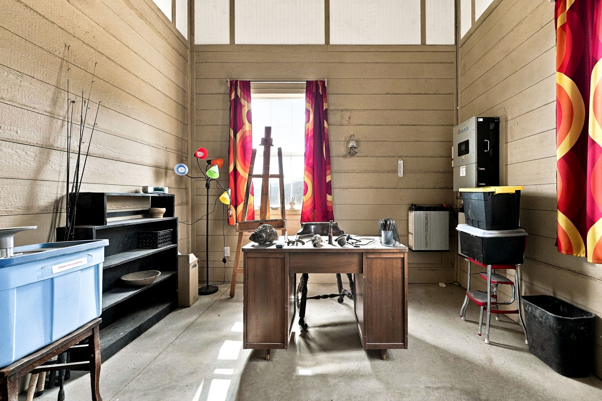 This rustic, multi-purpose workspace features wood-paneled walls and a concrete floor, creating a utilitarian yet creative atmosphere. The room is furnished with a central wooden desk, an artist's easel positioned by a window with vibrant patterned curtains, and open black shelving for storage. A modern power storage unit is mounted on the wall, and the space is illuminated by natural light streaming through the window and a colorful multi-bulb floor lamp.