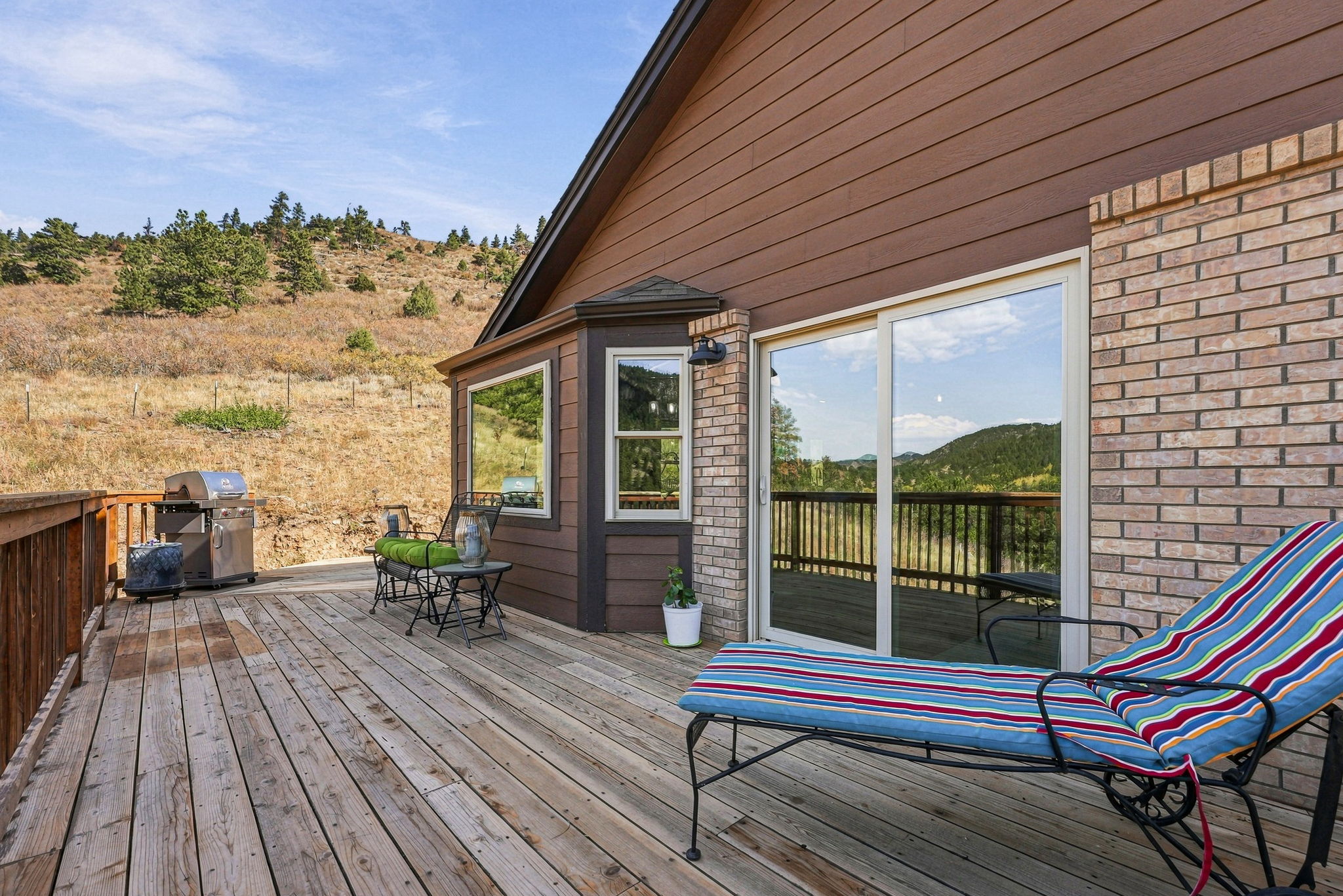 This spacious wooden deck offers a scenic outdoor living space with a view of the surrounding hilly landscape. The area features a striped lounge chair, a small bistro set, and a stainless steel grill, all set against a home exterior finished with brown siding and brick accents. Large sliding glass doors provide a seamless transition between the indoor and outdoor spaces, creating an inviting atmosphere for relaxation.