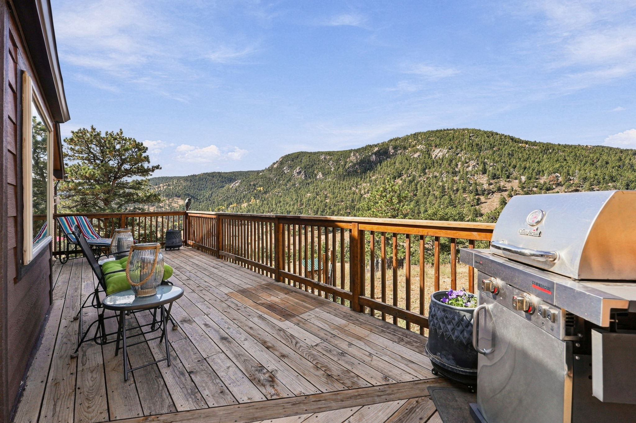 This spacious wooden deck offers a panoramic view of a lush, tree-covered mountain landscape under a clear blue sky. The area is furnished with a small bistro table, chairs with green cushions, and a stainless steel grill, making it an ideal spot for outdoor dining and relaxation. The deck features a sturdy wooden railing and provides a serene, elevated vantage point for enjoying the natural surroundings.