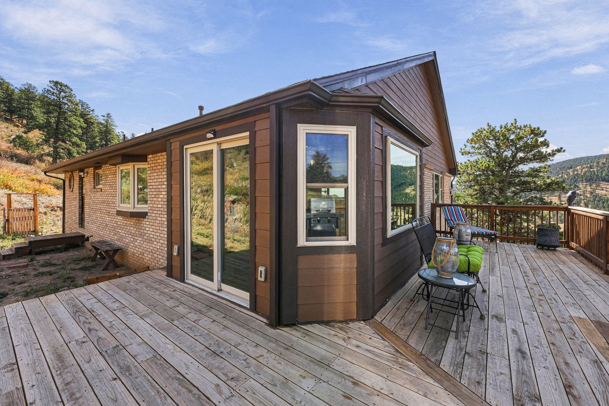 This image captures a spacious wooden deck attached to a home with a mix of brick and dark-toned siding. The deck features outdoor seating, including chairs with bright green cushions, and offers expansive views of the surrounding mountainous landscape. A sliding glass door provides access to the interior, while the elevated position emphasizes the home's scenic, rural setting.