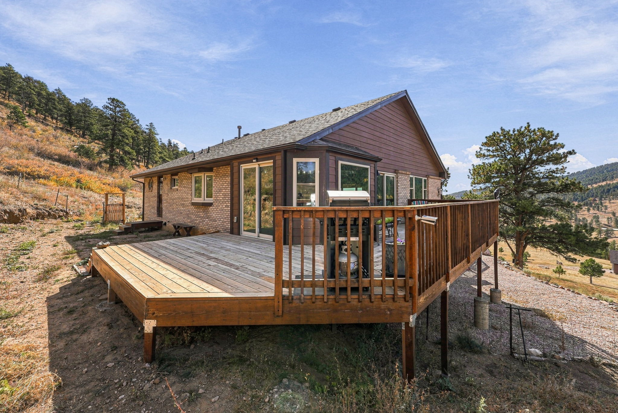 This image captures a spacious, elevated wooden deck attached to the rear of a home, offering expansive views of a hilly, natural landscape. The house features a combination of light-colored brick and dark horizontal siding, with large windows and sliding glass doors providing access to the outdoor living space. A gas grill is positioned on the deck, which is enclosed by a wooden railing, creating a perfect vantage point for enjoying the surrounding mountainous terrain.