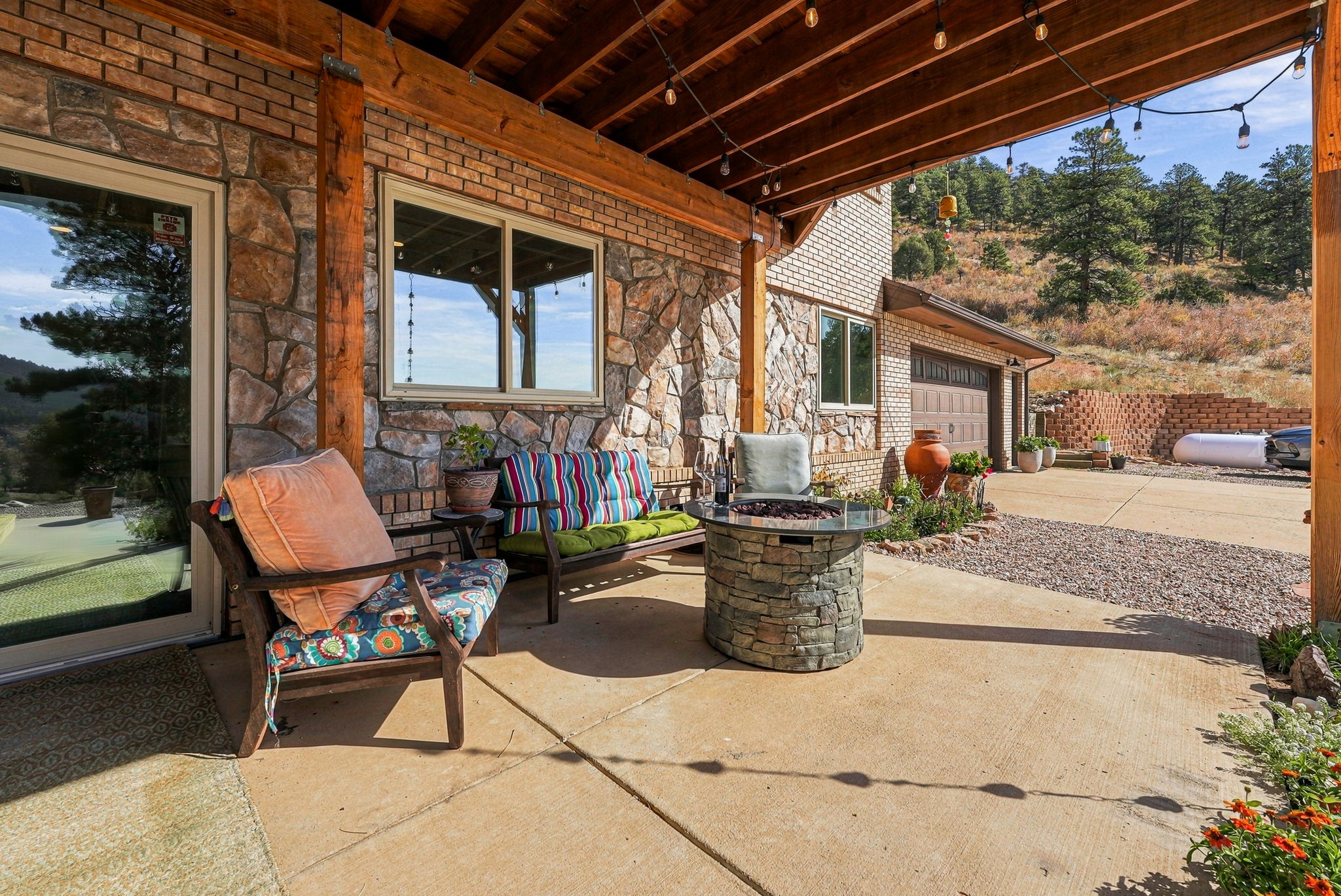 This inviting covered patio features a rustic stone-clad wall and a warm wood-beamed ceiling adorned with string lights. The space is furnished with a comfortable armchair, a colorful striped bench, and a central stone fire pit, creating a cozy outdoor seating area. The concrete patio overlooks a scenic, hilly landscape with mature trees, offering a peaceful and private atmosphere.