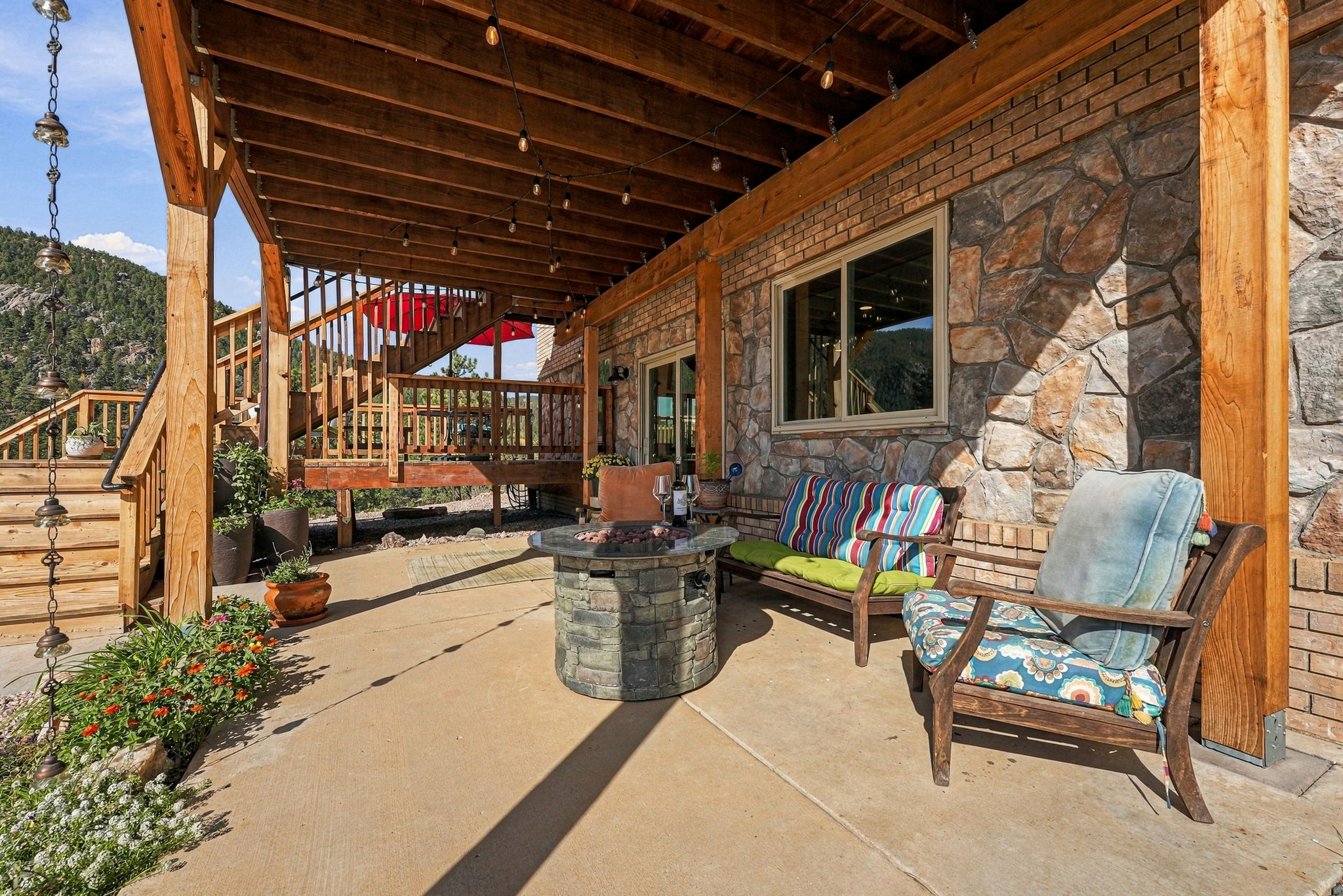 This inviting covered patio features a rustic stone-clad wall and exposed wooden ceiling beams, creating a warm, mountain-inspired atmosphere. The space is furnished with a stone-topped fire pit, a wooden bench with colorful striped cushions, and a matching armchair, all set against a backdrop of natural landscape. A wooden staircase leads up to an elevated deck, while string lights overhead add a cozy glow to this outdoor living area.