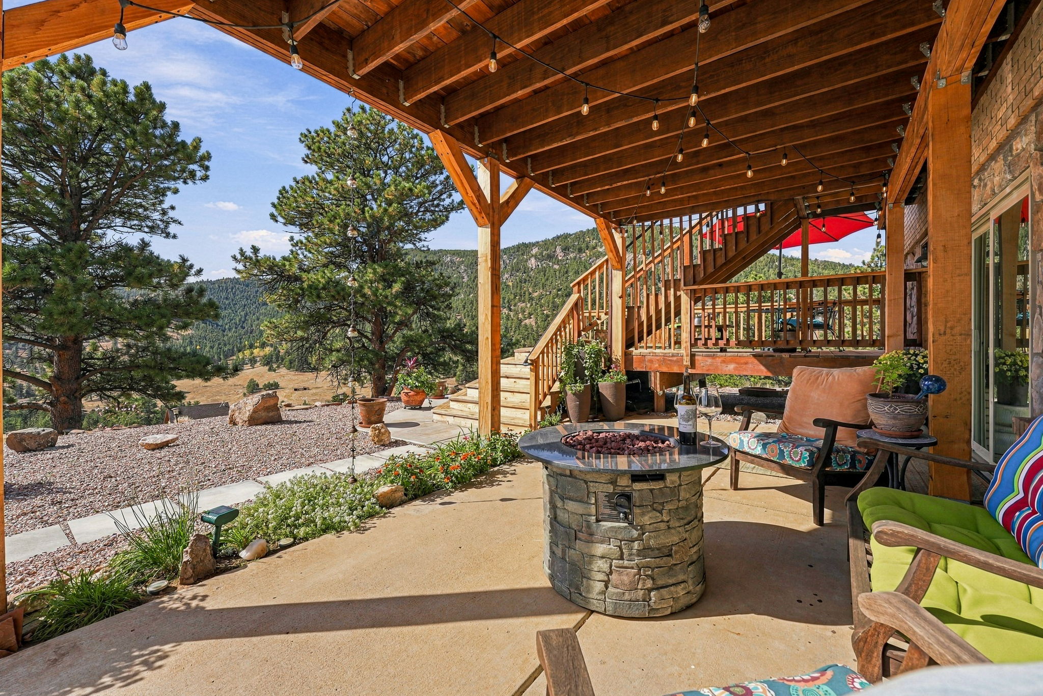 This inviting covered patio features a stone-clad fire pit as a central gathering point, complemented by comfortable outdoor seating. The space is sheltered by a rustic timber-framed structure with overhead string lighting, creating a warm atmosphere that overlooks a scenic, mountainous landscape. A wooden staircase leads up to an elevated deck, seamlessly blending indoor-outdoor living with expansive natural views.