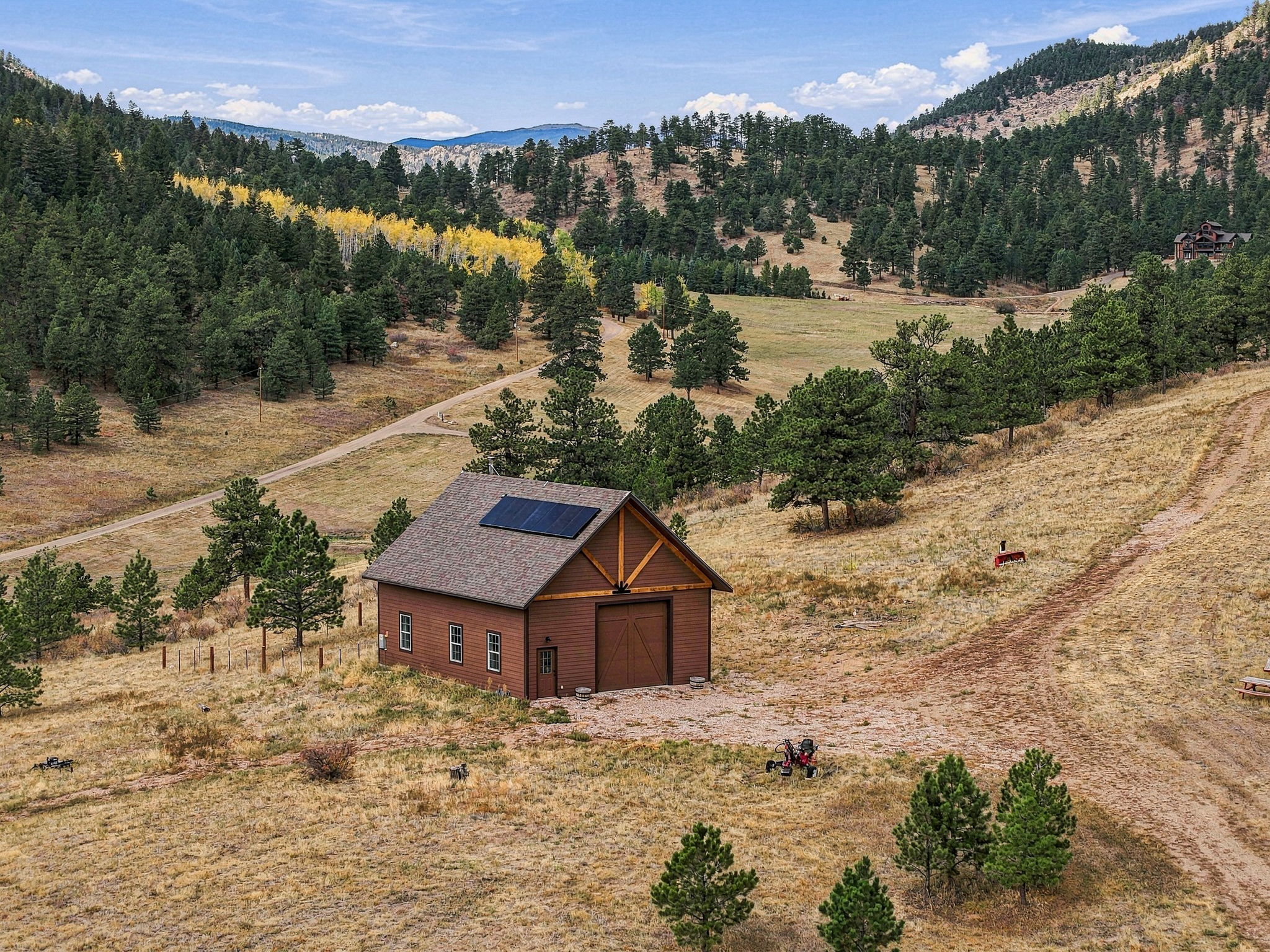 This aerial perspective captures a rustic, brown-sided barn-style structure nestled within a sprawling, mountainous landscape. The building features a gabled roof equipped with solar panels, a large central garage door, and a decorative timber truss detail above the entrance. The surrounding property is characterized by rolling hills, patches of evergreen and deciduous trees, and a dirt access road, creating a serene and secluded rural atmosphere.