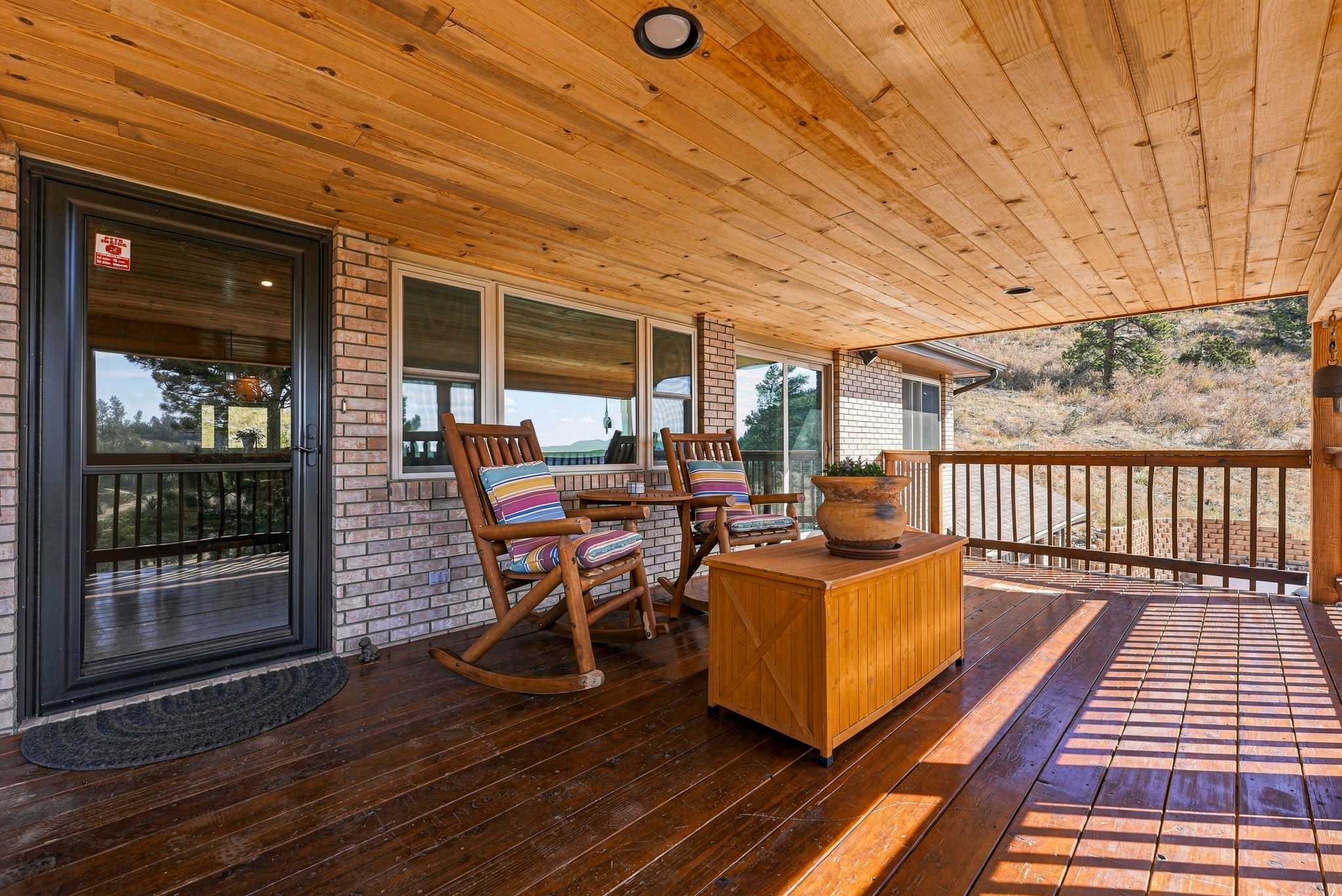 This spacious wooden deck features a covered area with a natural wood-paneled ceiling and a brick exterior wall. Two wooden rocking chairs with colorful striped cushions are positioned near a central wooden storage chest topped with a decorative planter. The deck overlooks a scenic, hilly landscape, providing a peaceful outdoor living space.