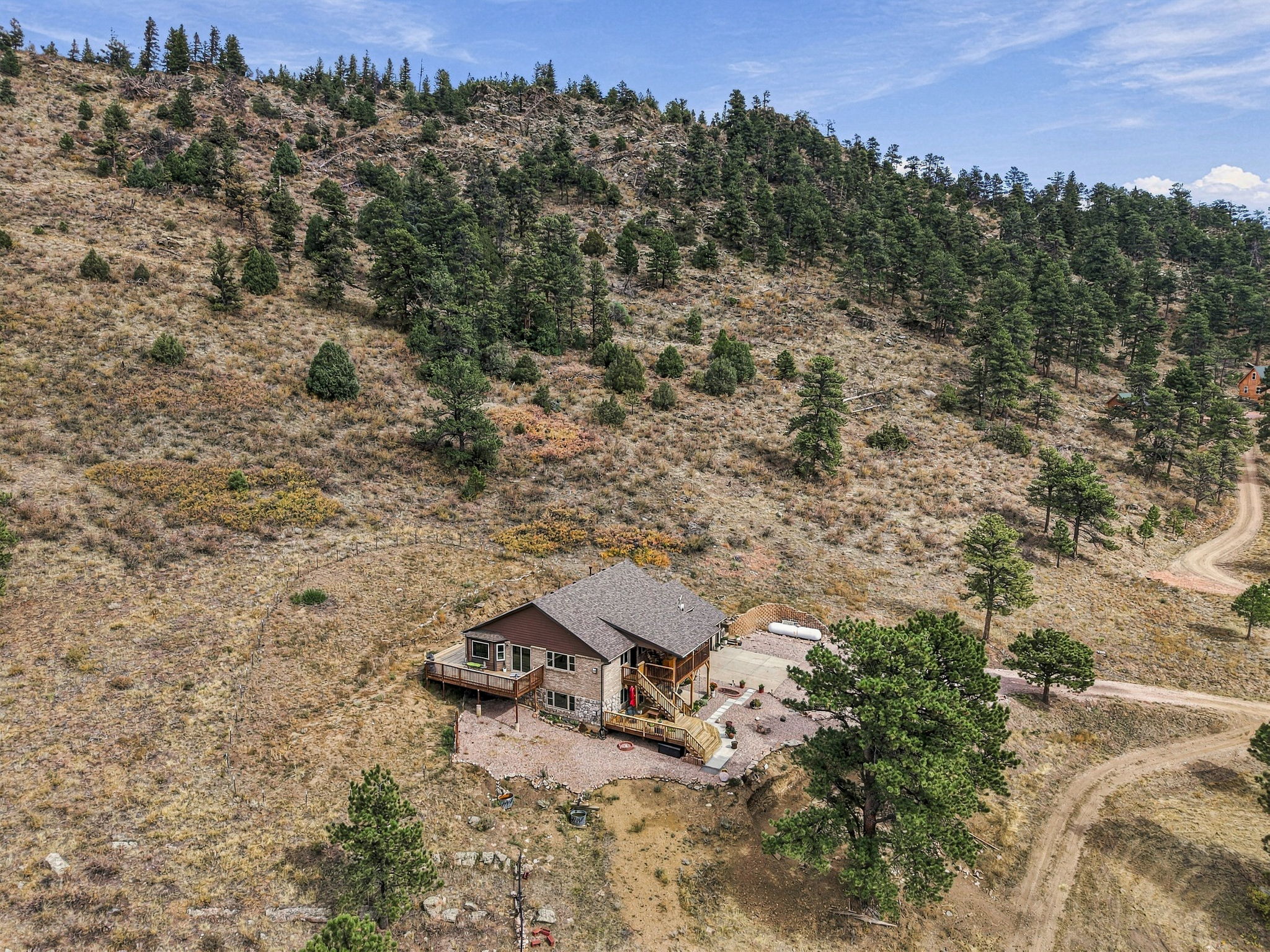 This high-angle aerial shot captures a multi-level home nestled into a sloping, rugged hillside landscape. The house features a combination of brown siding and stone accents, complemented by expansive wooden decks and a gravel driveway. The surrounding terrain is characterized by dry, grassy slopes dotted with evergreen trees, creating a secluded and natural mountain setting.