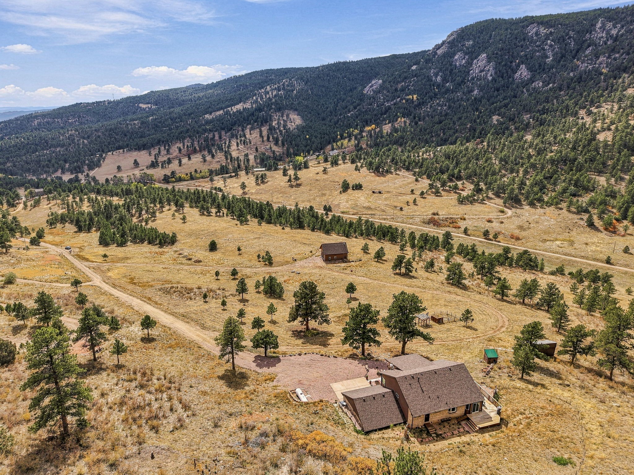 This high-angle aerial view captures a sprawling rural property nestled against a vast, forested mountain range. The main residence features a multi-gabled roofline and a wooden deck, situated on a large, dry grassy lot dotted with scattered pine trees and a secondary outbuilding. A winding dirt driveway leads up to the home, emphasizing the secluded and expansive nature of this mountain landscape.