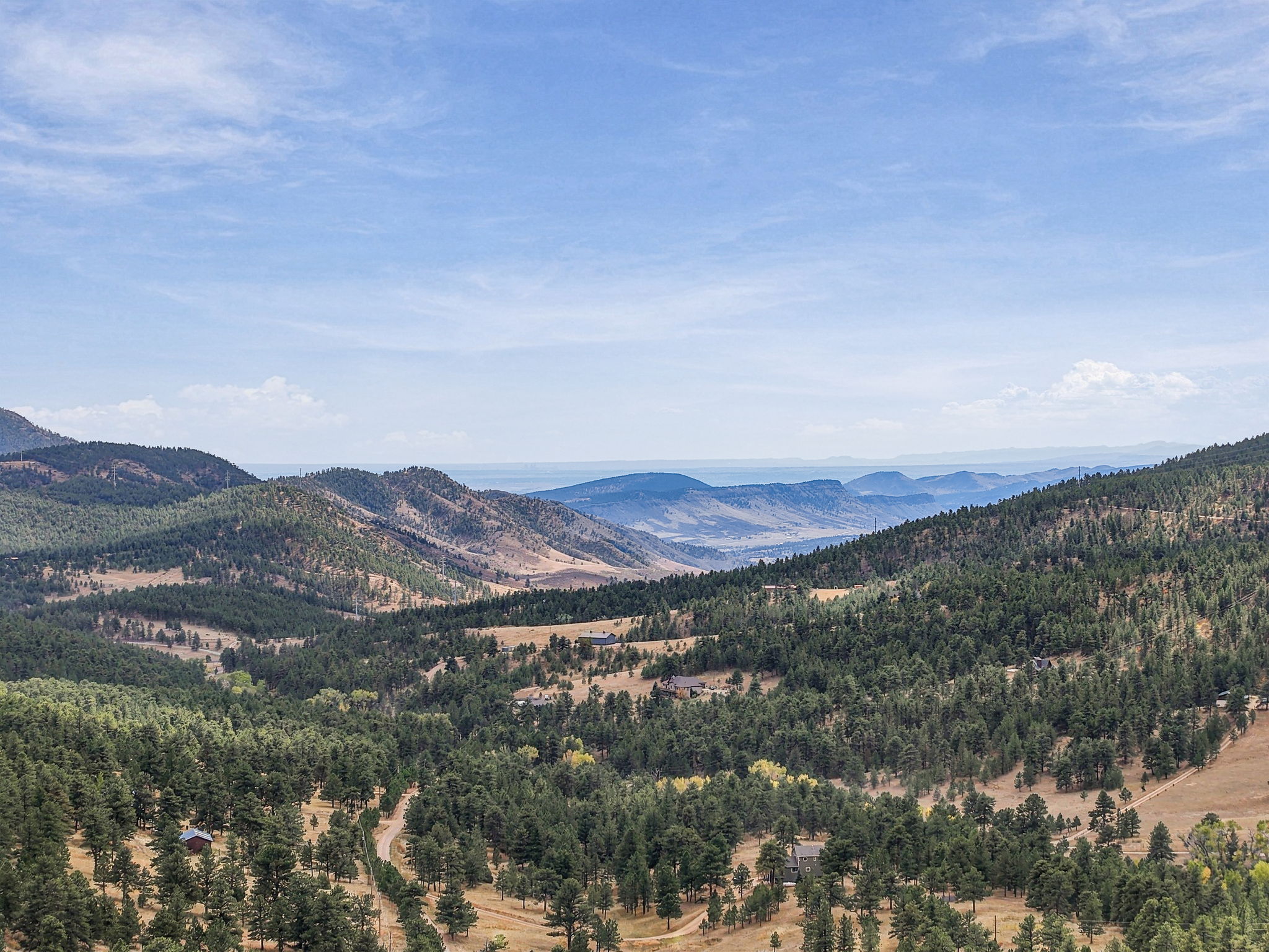 This expansive aerial view captures a vast, mountainous landscape characterized by rolling hills covered in dense pine forests and patches of dry, golden grassland. Several secluded residential homes are nestled within the terrain, connected by winding dirt roads that traverse the rugged topography. The scene offers a serene, panoramic perspective of a remote, natural setting under a bright, expansive sky.