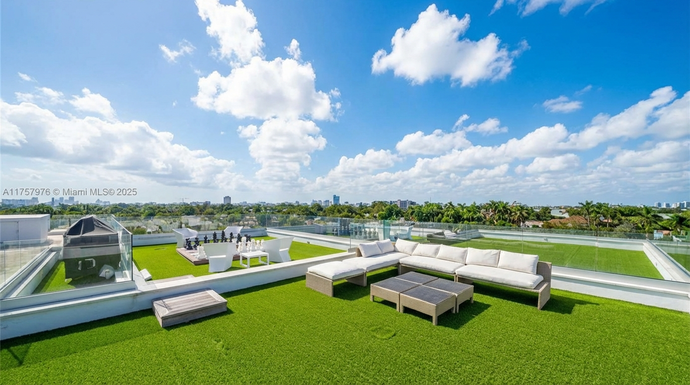 This image showcases a luxurious rooftop patio or deck, featuring artificial turf and stylish outdoor furniture. The space includes a seating area with a large sectional sofa, coffee tables, and a dining area. The clear glass railings offer unobstructed panoramic views of the cityscape and lush greenery, adding to the property's appeal for outdoor entertaining and relaxation.