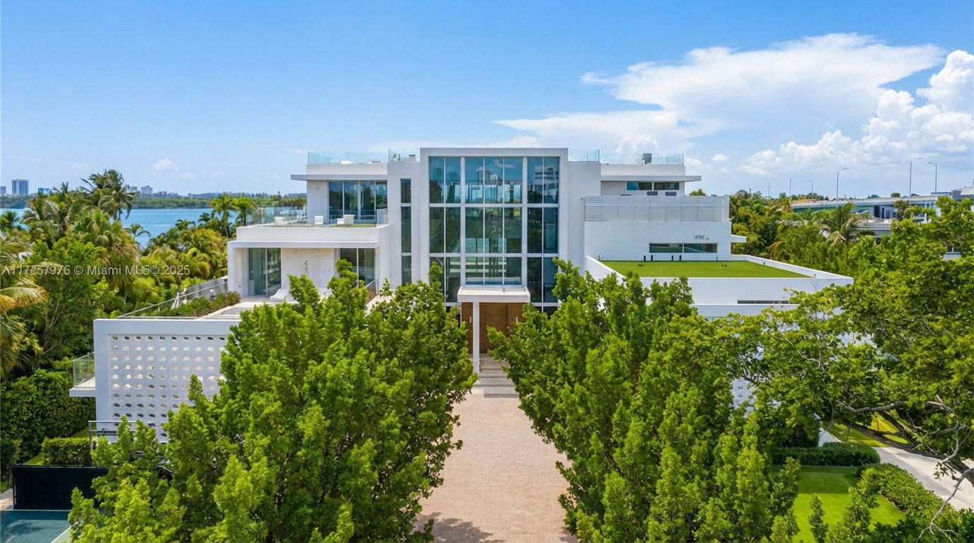 This is an aerial view of a modern luxury home showcasing its impressive architectural design. The residence features clean lines, a flat roof with a green roof section, extensive glass windows, and multiple levels. The property is surrounded by lush green trees and landscaping, enhancing its privacy and curb appeal.
