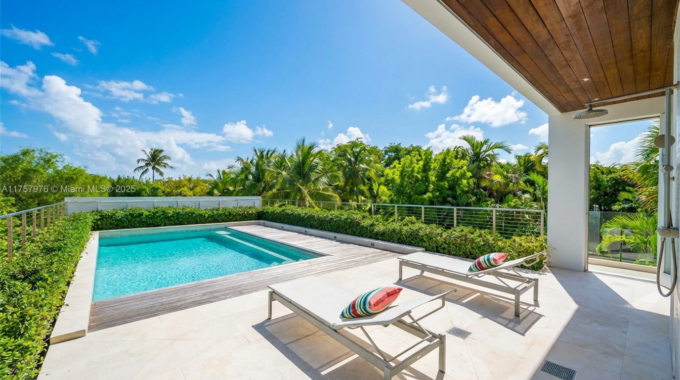 This image showcases a modern outdoor pool area. The pool is surrounded by a wooden deck and patio with white tile. Two lounge chairs with colorful striped pillows face the pool, creating inviting seating arrangement. Lush greenery and trees provide privacy and a tranquil atmosphere.