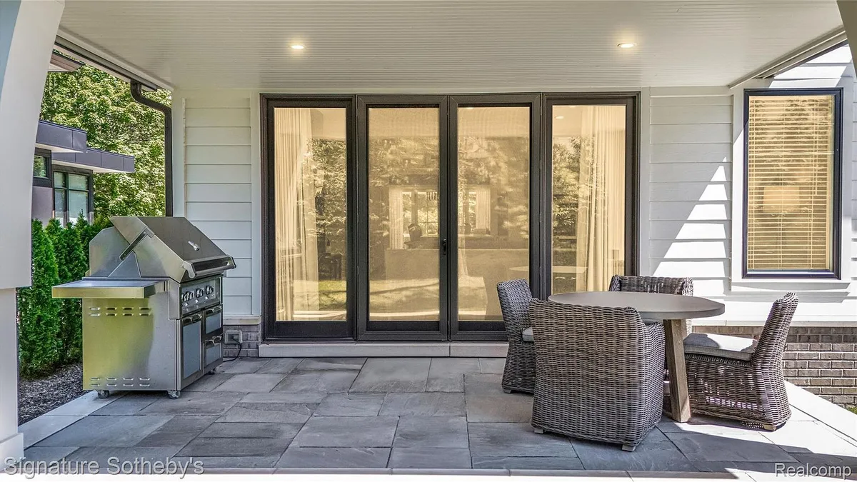 This image showcases an outdoor patio area, perfect for relaxation and entertaining. Key features include a built-in barbecue grill, a round dining table, and comfortable wicker chairs. The patio is paved with gray stone, creating a sophisticated and inviting space, while large glass doors create a seamless transition to the home's interior.