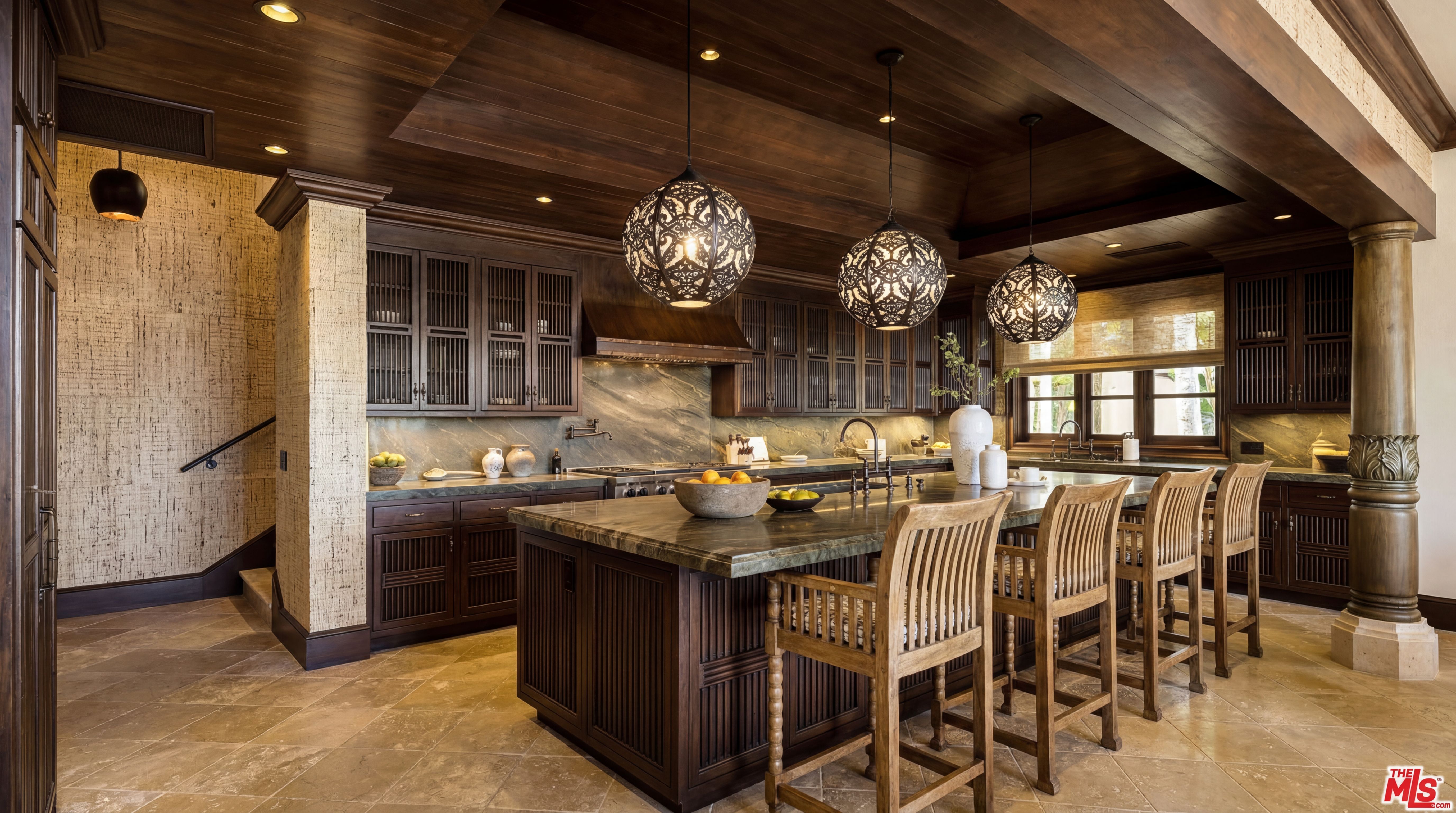 This luxurious, high-end kitchen features rich, dark wood cabinetry and a spacious island topped with custom natural stone. The sophisticated design is accented by unique, ornate pendant lighting, intricate woodwork, and designer wallpaper, creating an opulent and warm atmosphere. A large, well-lit island provides ample workspace and seating, making it an ideal focal point for both cooking and entertaining.