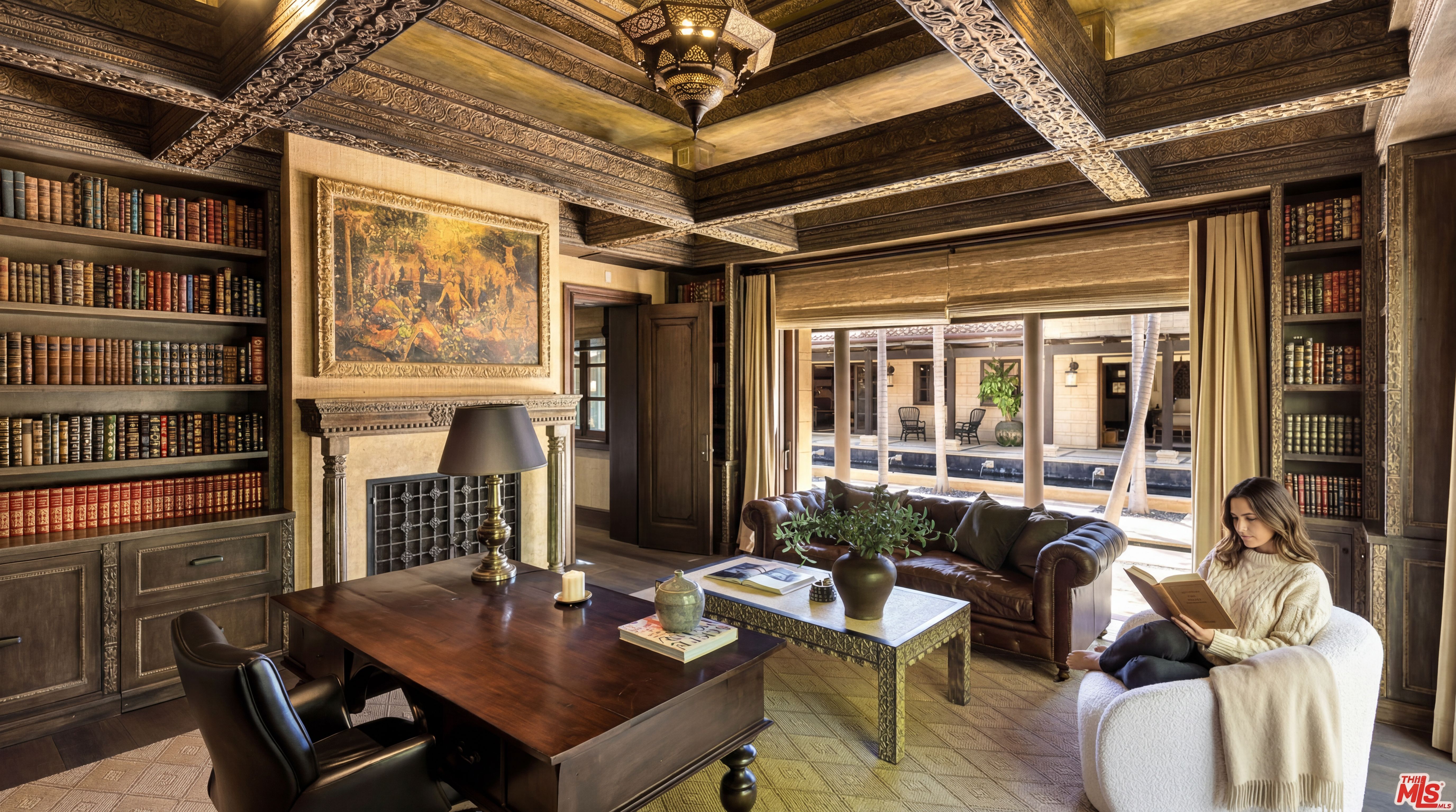This elegant home office features rich wood paneled walls, ornate ceiling detailing, and built-in bookshelves stocked with vintage books. The space is anchored by a solid wood desk, a leather sofa, and a comfortable reading chair, all opening up to a bright, serene courtyard view through glass doors. The warm lighting and sophisticated decor create a refined, scholarly atmosphere.
