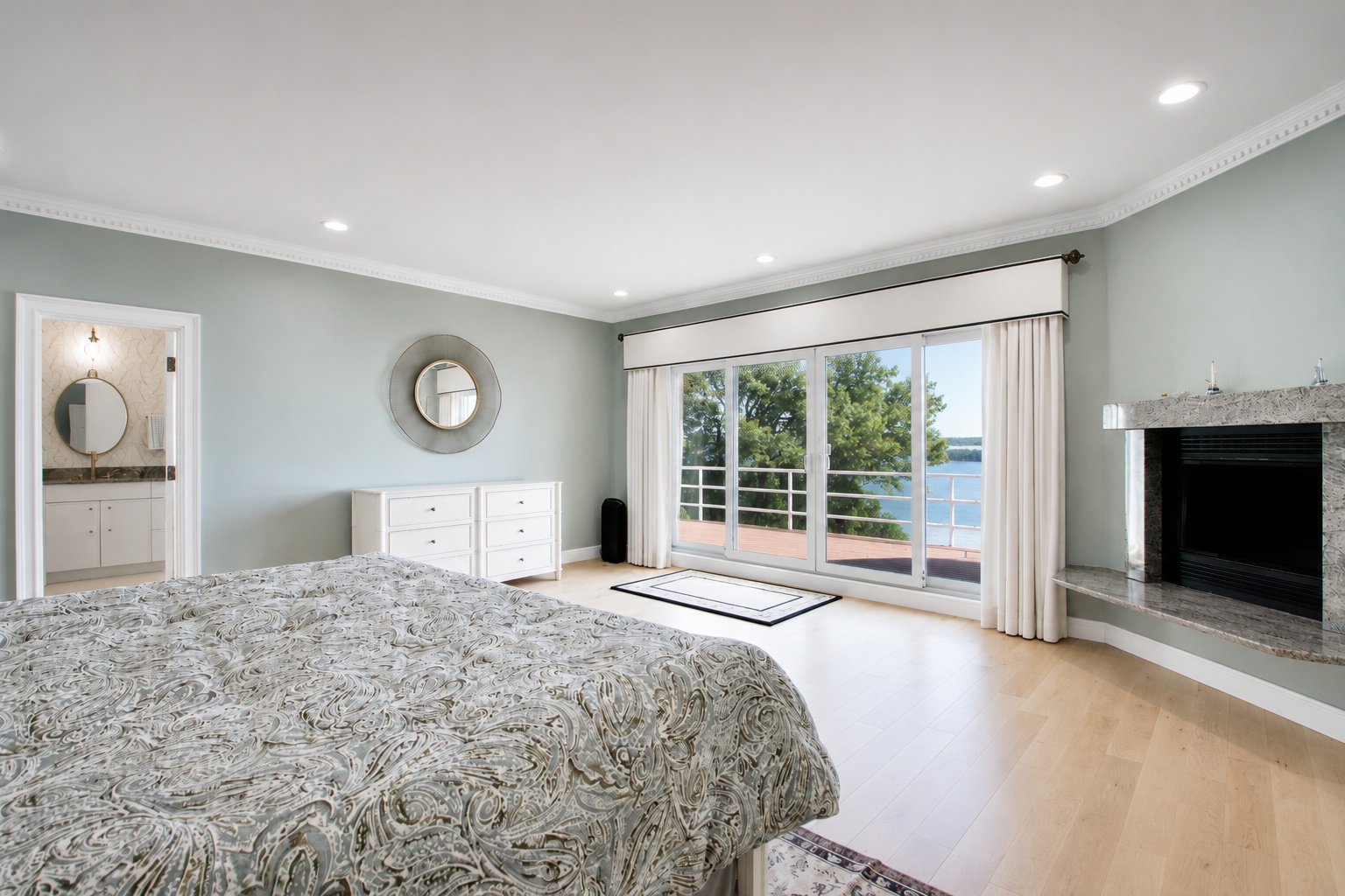 This spacious primary bedroom features soft blue-gray walls, light hardwood flooring, and a large fireplace with a granite surround. A set of floor-to-ceiling sliding glass doors provides expansive views of the outdoors and natural light, complemented by a white dresser and an elegant round mirror. The room offers a serene, airy atmosphere that flows seamlessly into the attached bathroom.