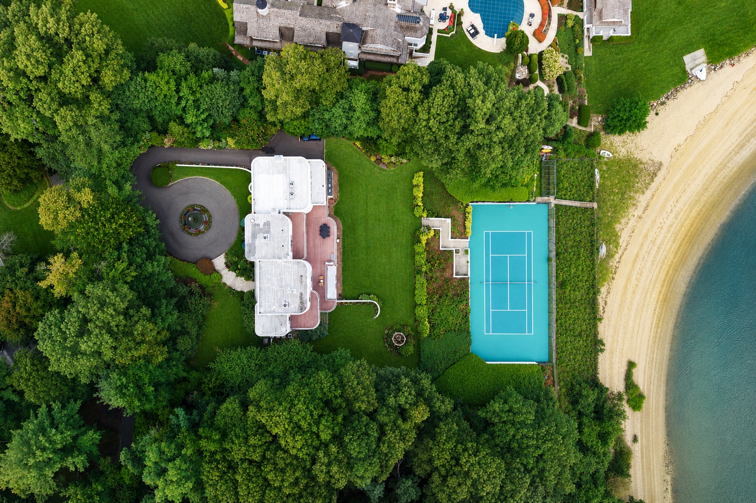This stunning aerial view captures a sprawling waterfront estate characterized by a large, modernist mansion nestled among extensive, mature woodlands. The property boasts impressive luxury amenities, including a private, vibrant blue tennis court and a circular driveway feature. The seamless transition from the manicured lawns to the sandy beach and serene waterfront emphasizes its elite, resort-like lifestyle.