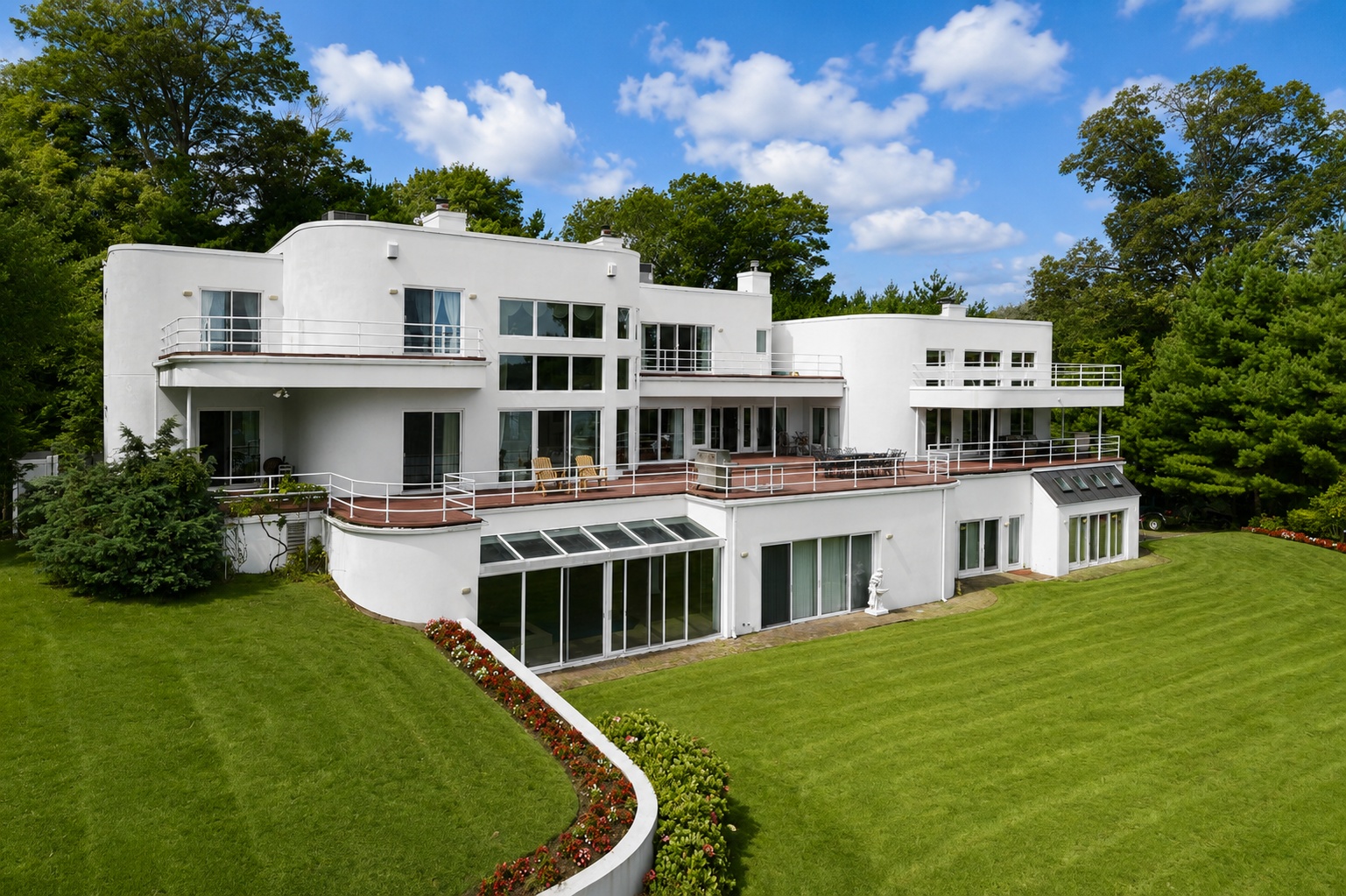 This striking multi-level residence features a distinct Art Deco-influenced curved architectural style with a pristine white exterior. The property boasts an expansive elevated deck, large glass sliding doors, and a greenhouse-style sunroom, all overlooking a beautifully manicured lawn and lush surrounding greenery. Its unique design creates an impressive, airy aesthetic that seamlessly integrates indoor and outdoor living spaces.