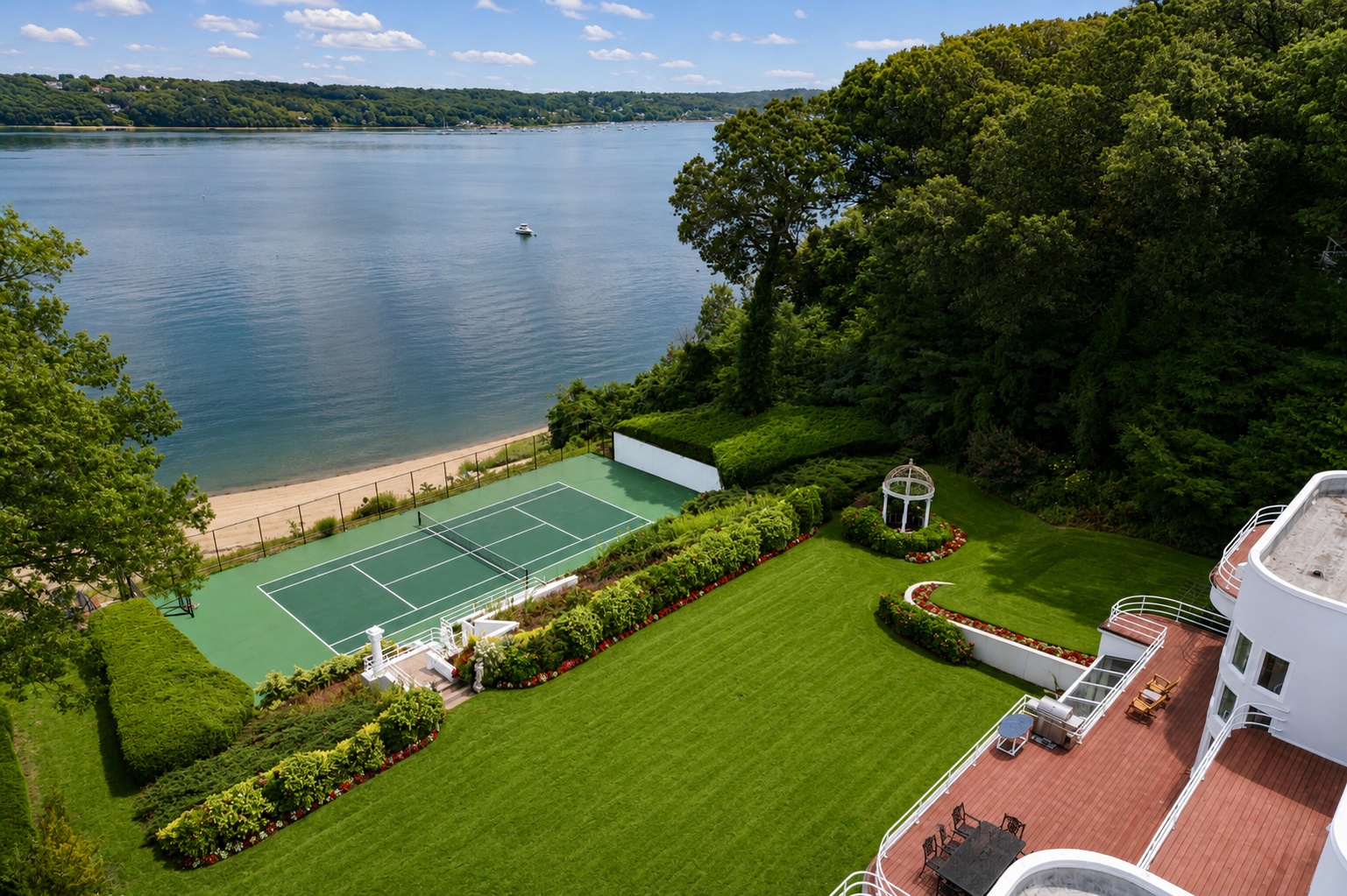 This stunning high-angle aerial view captures a luxurious waterfront estate, featuring a vast, meticulously landscaped green lawn that leads directly to a private tennis court and sandy beach. The property includes expansive multi-level wooden decking directly attached to a contemporary white home, a charming white gazebo, and lush surrounding woodland. The serene water view and highly manicured grounds create a tranquil and prestigious coastal atmosphere.
