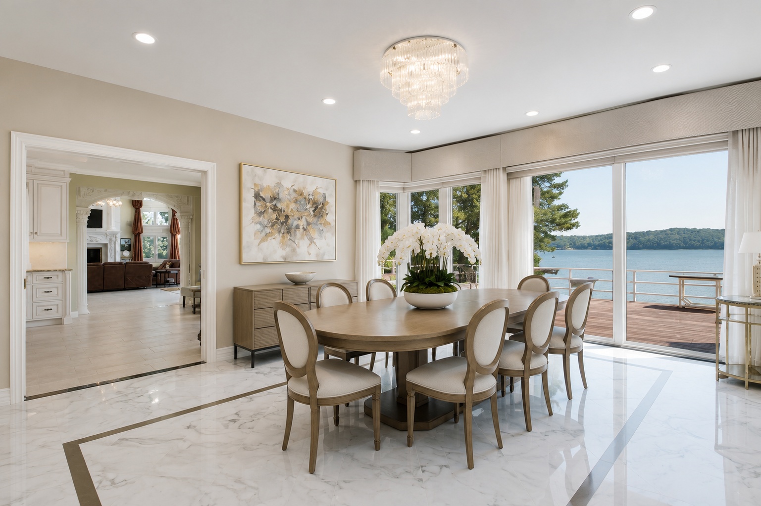 This elegant dining room features a large oval wooden table surrounded by light-toned upholstered chairs, set beneath a stunning tiered crystal chandelier. The space boasts polished marble flooring with decorative inlay and expansive sliding glass doors that provide seamless indoor-outdoor living with panoramic lake views. A chic sideboard and abstract artwork complete the sophisticated, bright, and airy atmosphere.