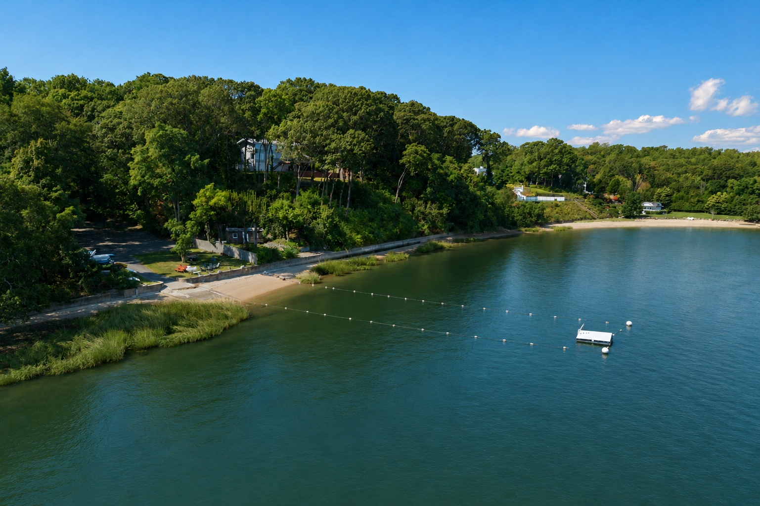 This aerial view showcases a waterfront property nestled along a heavily wooded hillside overlooking a calm bay. The site features a sandy beach area, a concrete bulkhead, and a floating swim platform securely tethered in the water, offering a serene and private recreational setting.
