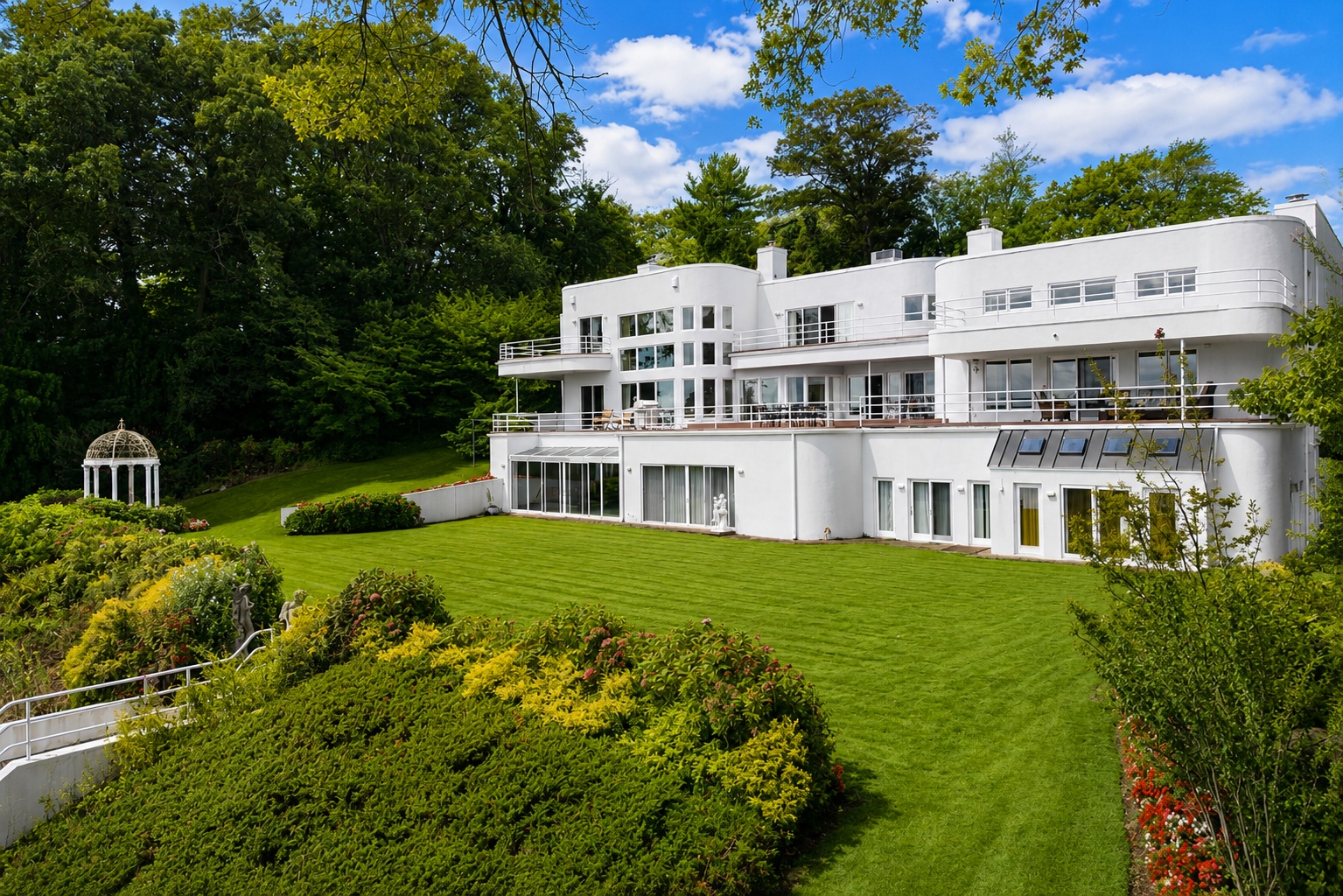This spacious, modern white residence features a striking curvilinear architectural design and multiple levels of expansive balconies that maximize outdoor living space. The property is set against a backdrop of mature woodland and includes a meticulously manicured lawn, tiered landscaping, and a classic garden gazebo. The overall aesthetic is one of sophisticated luxury, blending grand architectural forms with harmonious natural beauty.