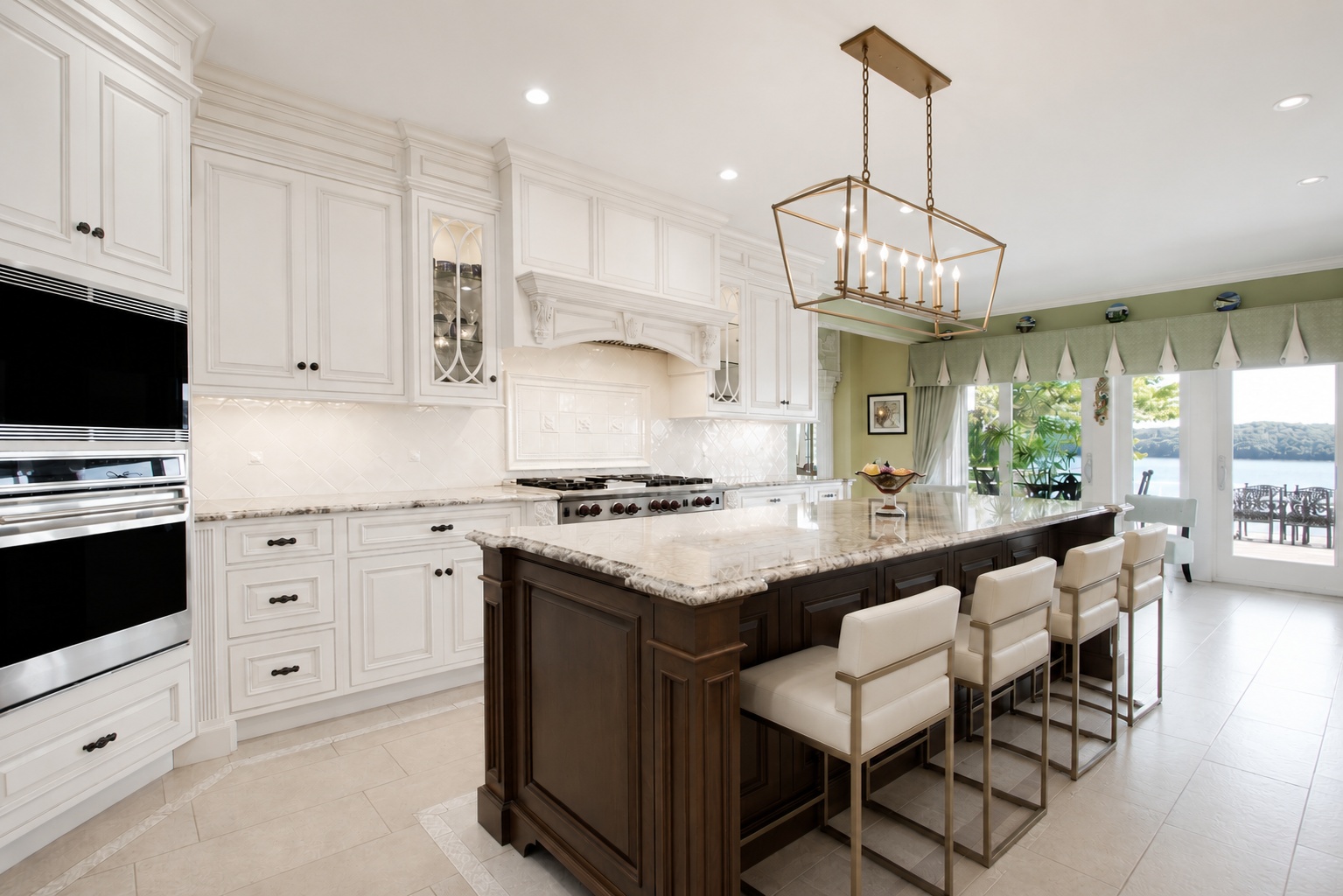 This elegant and spacious kitchen features a prominent dark wood center island accented with contemporary cream-colored bar stools and a luxurious stone countertop. The perimeter is defined by classic white cabinetry, high-end stainless steel appliances, and sleek, light-colored tile flooring. Large glass patio doors at the rear provide an abundance of natural light and serene water views, creating an inviting and sophisticated living space.