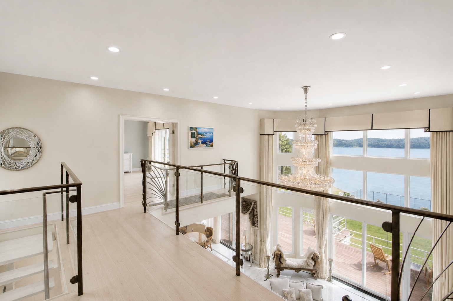 This elegant upper-level mezzanine features light-toned hardwood flooring and a stylish metal and glass railing system that overlooks the main living area. A stunning multi-tiered crystal chandelier hangs as a central focal point, while floor-to-ceiling windows provide expansive, picturesque views of the lake. The decor is refined and airy, suggesting a luxurious home with a modern architectural sensibility.