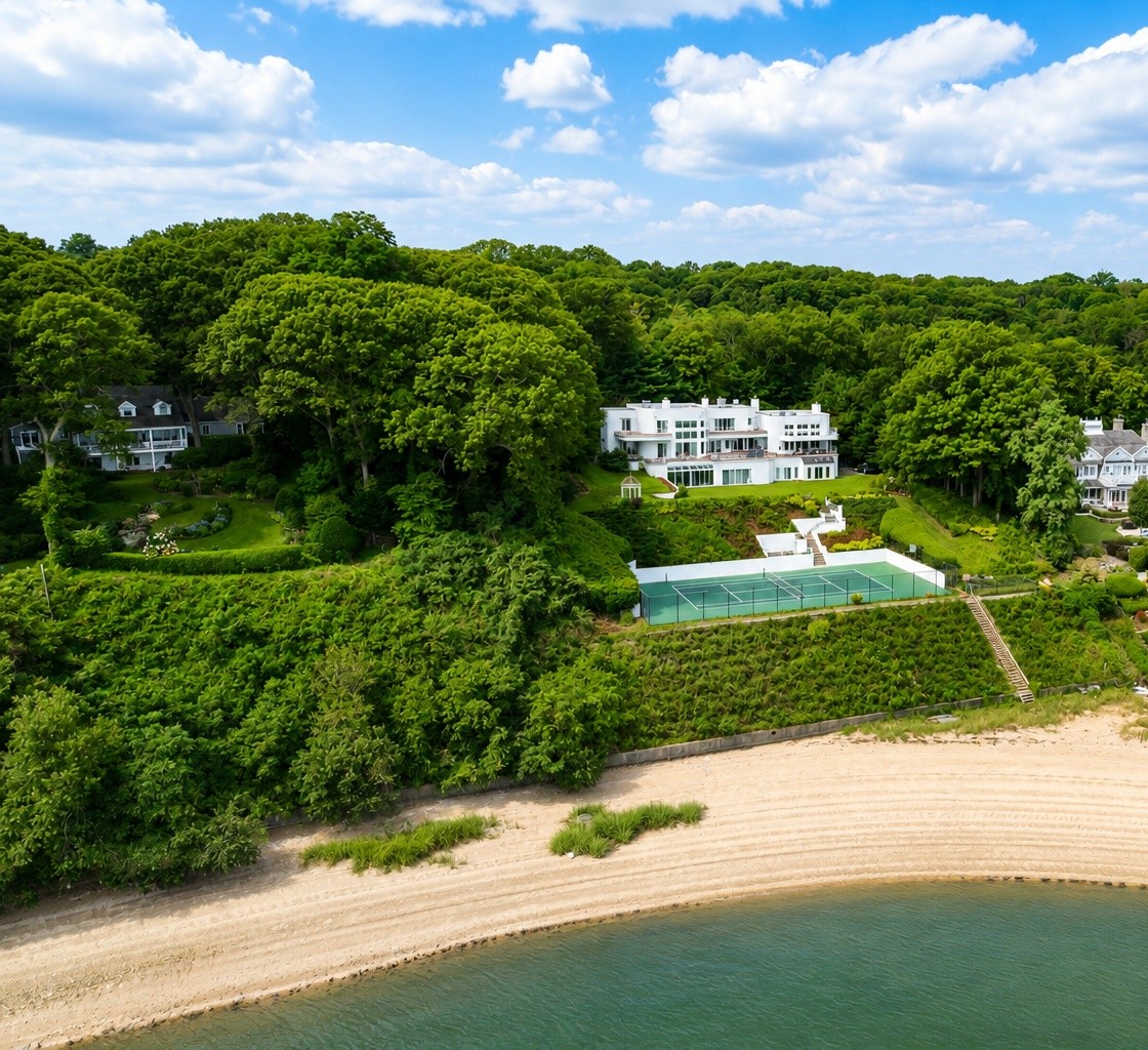 This aerial view showcases a luxurious waterfront estate, featuring a prominent white modern villa nestled against a dense, lush hillside. The property includes a private tennis court situated on a terraced slope above the beach, offering a peaceful and exclusive atmosphere. The expansive green surroundings and direct beach access highlight the premier coastal living experience this home provides.