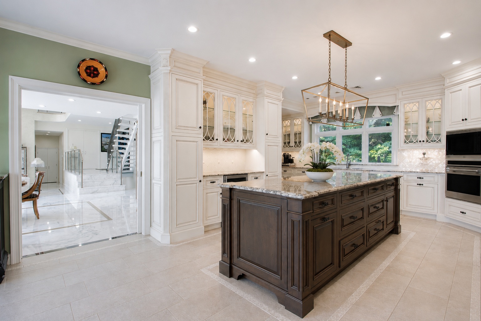 This elegant and spacious kitchen features a rich, dark-stained wood island topped with stone countertops, acting as a dramatic focal point against the surrounding white cabinetry. Notable features include a large, open floor plan that flows into an adjoining hallway with marble flooring and a statement staircase, creating a sophisticated and luxurious atmosphere. High-end finishes, such as a custom geometric chandelier and custom glass-front cabinetry, enhance the upscale, traditional design aesthetic.