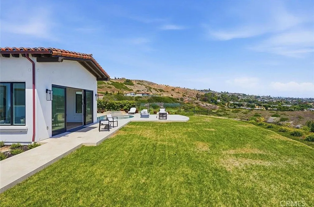 This image showcases the rear view of a modern house featuring a minimalist design with white stucco walls, dark-framed sliding glass doors, and a tiled roof edge. The home transitions to a well-maintained concrete patio area surrounding a small pool, which opens up to a vast, manicured lawn that overlooks rolling hills. The overall feel is one of serene luxury, emphasizing seamless indoor-outdoor living and scenic, panoramic landscape views.