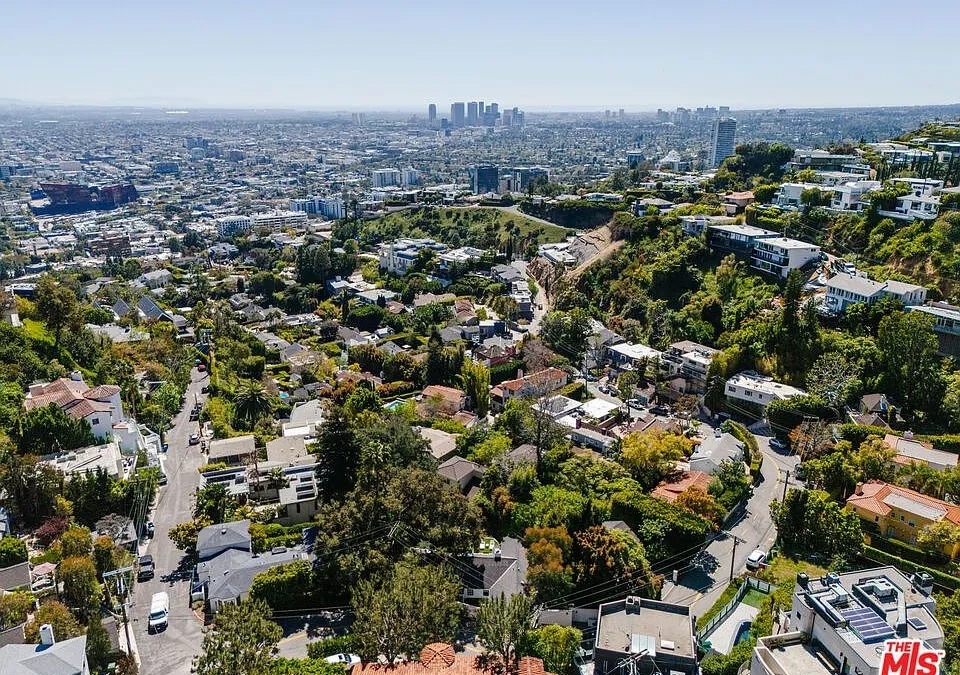 This aerial view showcases an expansive hillside neighborhood with various residential properties and abundant greenery. The backdrop offers a sweeping cityscape, suggesting a desirable location with scenic views. The image emphasizes the exclusive and elevated nature of the real estate.
