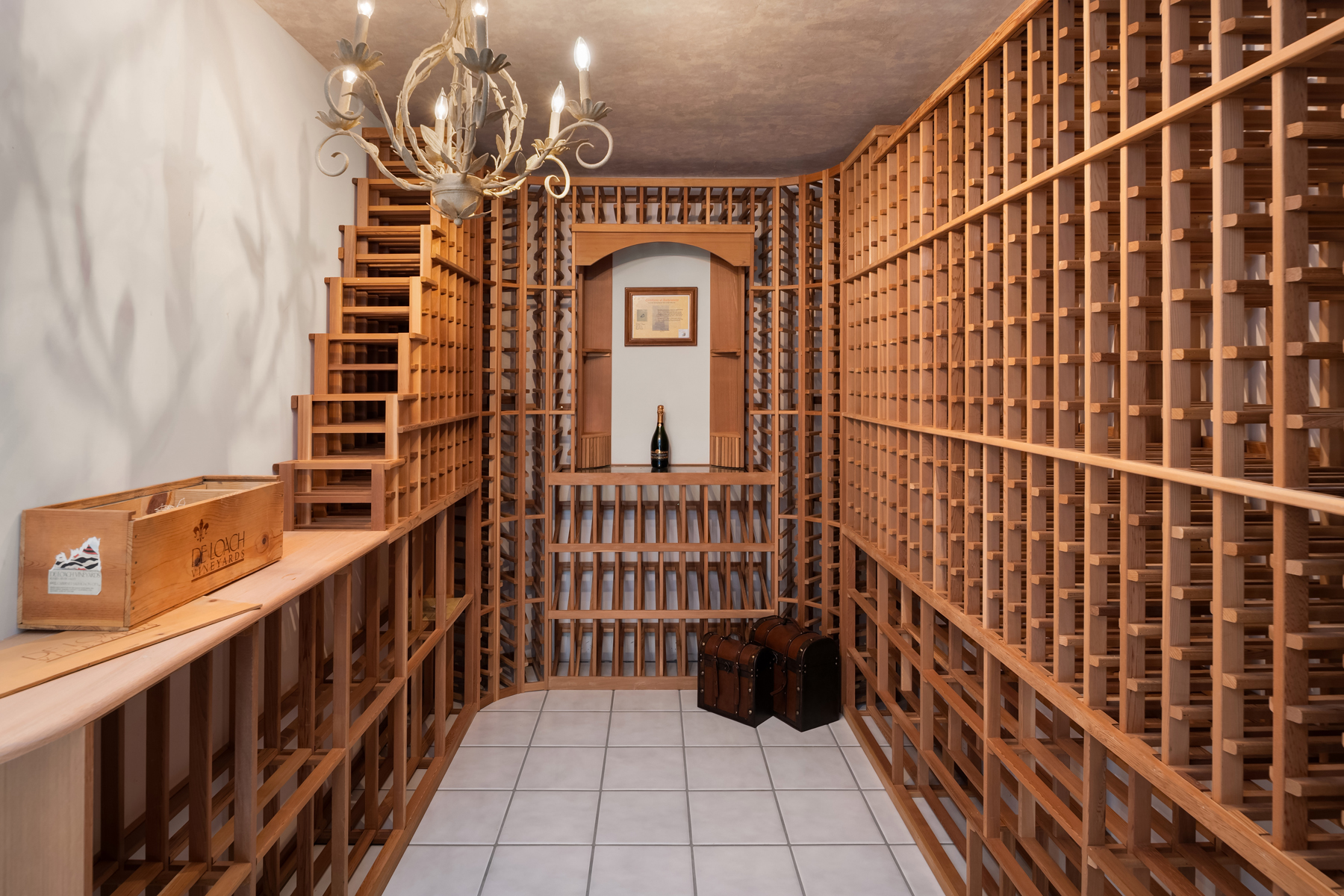 This is an interior shot of a well-organized wine cellar. The cellar features custom wooden racking, designed to hold numerous bottles of wine. A decorative chandelier hangs from the ceiling, and the floor is tiled. The overall impression is one of sophistication and careful preservation.