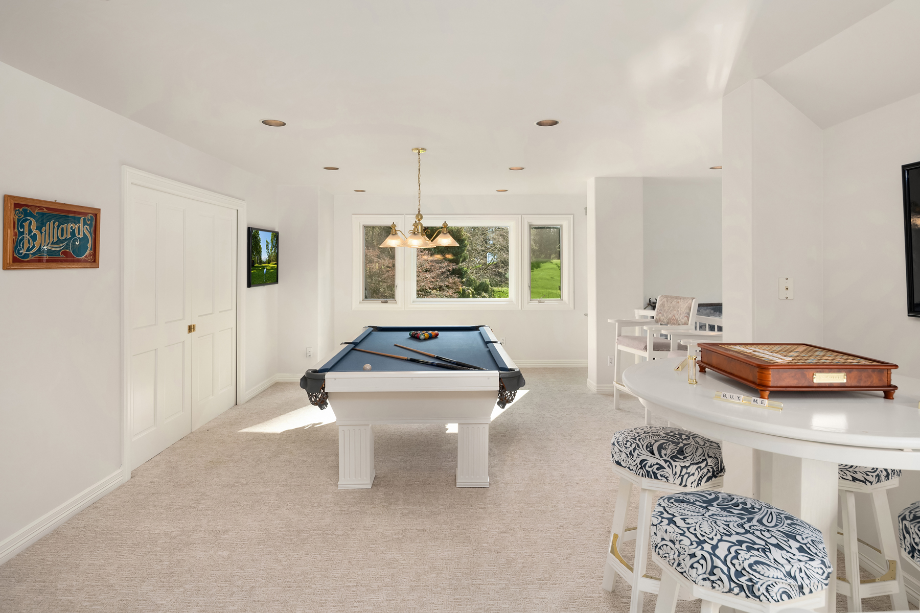 This is a well-lit basement featuring a billiards table with a blue felt top, positioned under a hanging light fixture and in front of a window. The room is painted in a clean white, complemented by light-colored carpeting, and includes a small bar area with stools and a wall-mounted television, creating a recreational space.