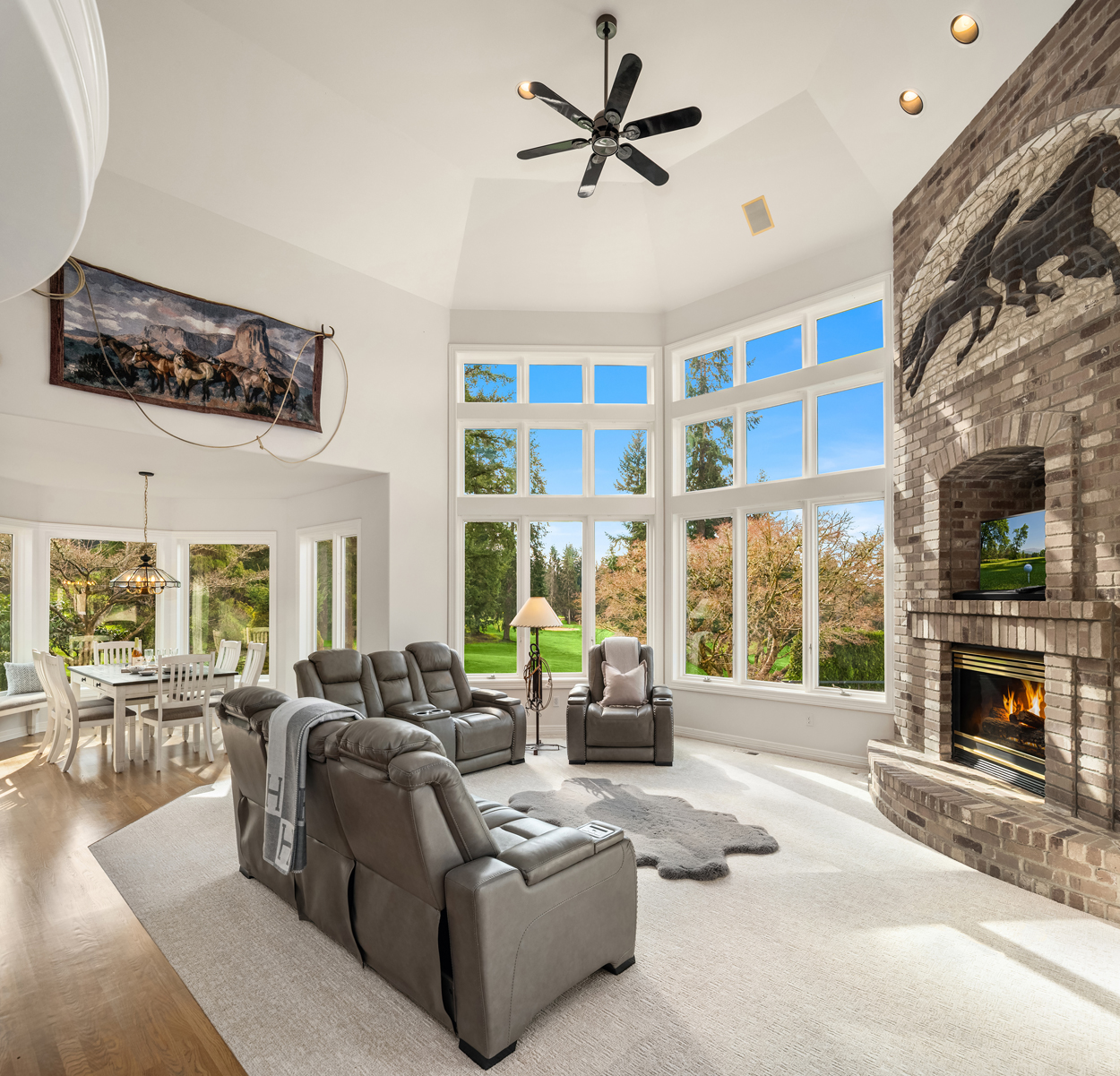 This is a spacious living room featuring a high vaulted ceiling, a large bay window offering abundant natural light and views of the outdoors, and a brick fireplace with a horse motif above. The room is furnished with comfortable gray leather seating and a neutral-toned rug, creating a welcoming and luxurious atmosphere. A dining area is visible in the background, suggesting an open-concept layout.