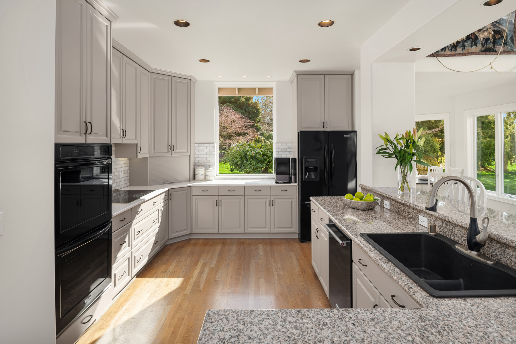 This is a well-lit kitchen featuring light gray cabinetry with black hardware, black appliances, and granite countertops. The kitchen has a window overlooking a garden, and the hardwood floors add warmth to the space. The overall impression is clean, modern, and inviting.
