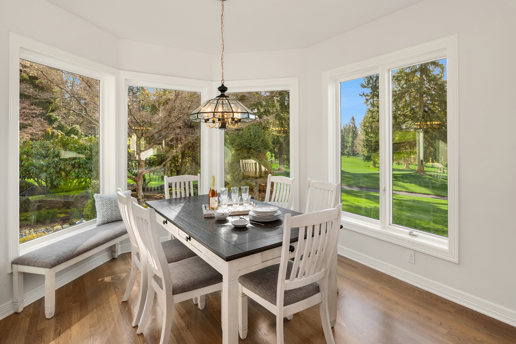 This is a bright and airy dining room featuring a dark-topped dining table with white legs and matching chairs with gray cushions. Large windows offer views of the surrounding greenery, and a stylish chandelier hangs above the table. A built-in bench seat is nestled under one of the windows, providing additional seating.