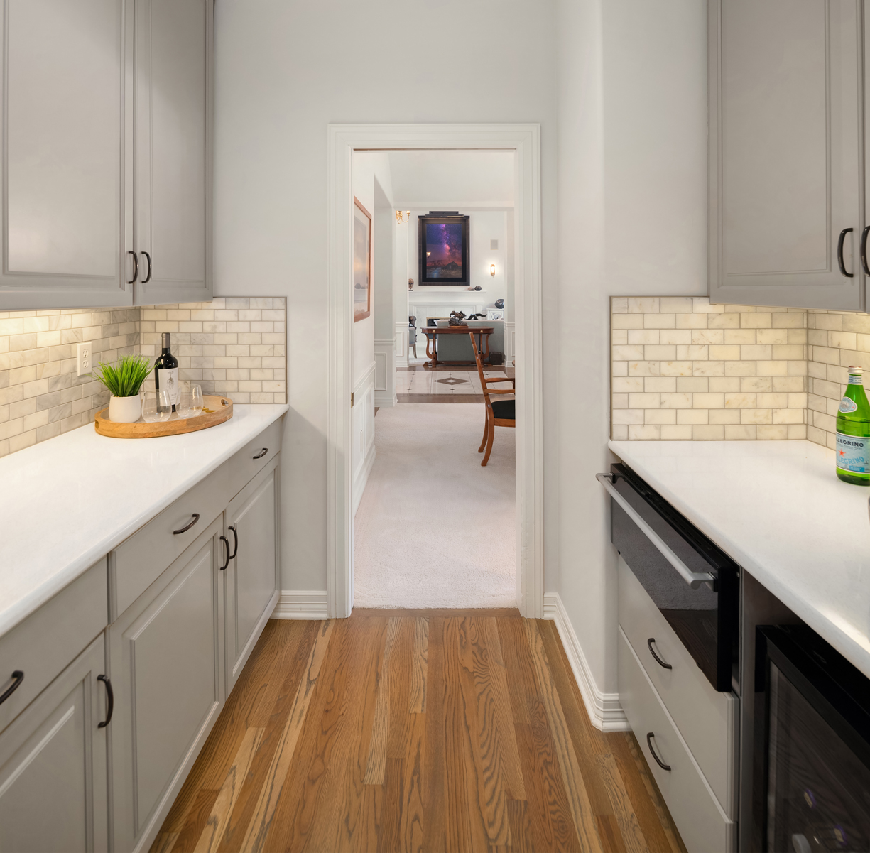 This interior shot showcases a hallway with light gray cabinetry on either side, featuring white countertops and a light marble backsplash. The warm wood flooring adds a touch of elegance, leading to a doorway that reveals a glimpse into a living area with a painting above a fireplace. The perspective is from a standing position, looking down the hallway.