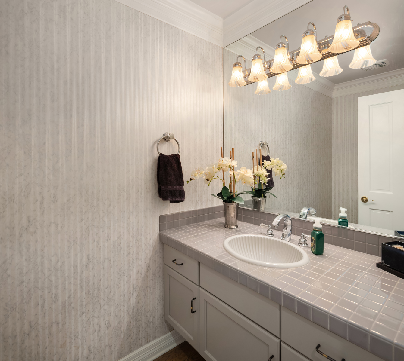 This is a well-maintained guest bathroom featuring a vanity with a white sink and tiled countertop. A large mirror with decorative lighting hangs above the sink, reflecting the room's neutral-toned wallpaper. A dark brown towel hangs on a towel ring to the left of the sink, and a vase of white flowers adds a touch of elegance.