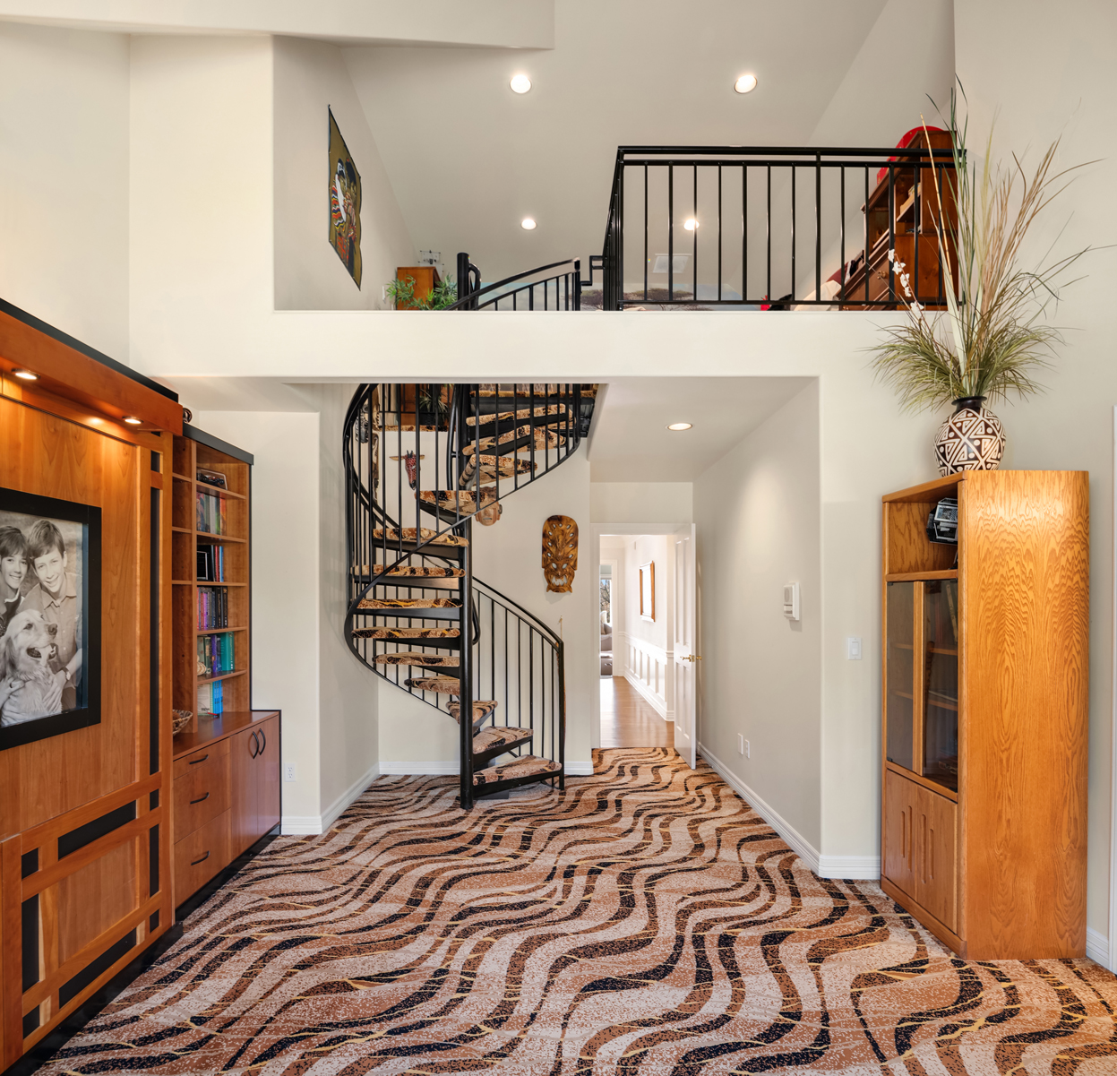 This interior shot showcases a grand hallway featuring a striking spiral staircase with black metal railings and patterned steps. The flooring has a unique wavy pattern in brown and beige tones, complemented by custom wooden built-in cabinetry on the left and a tall wooden cabinet on the right. The hallway leads to a bright, well-lit corridor, and a loft area is visible above, adding to the home's architectural interest.