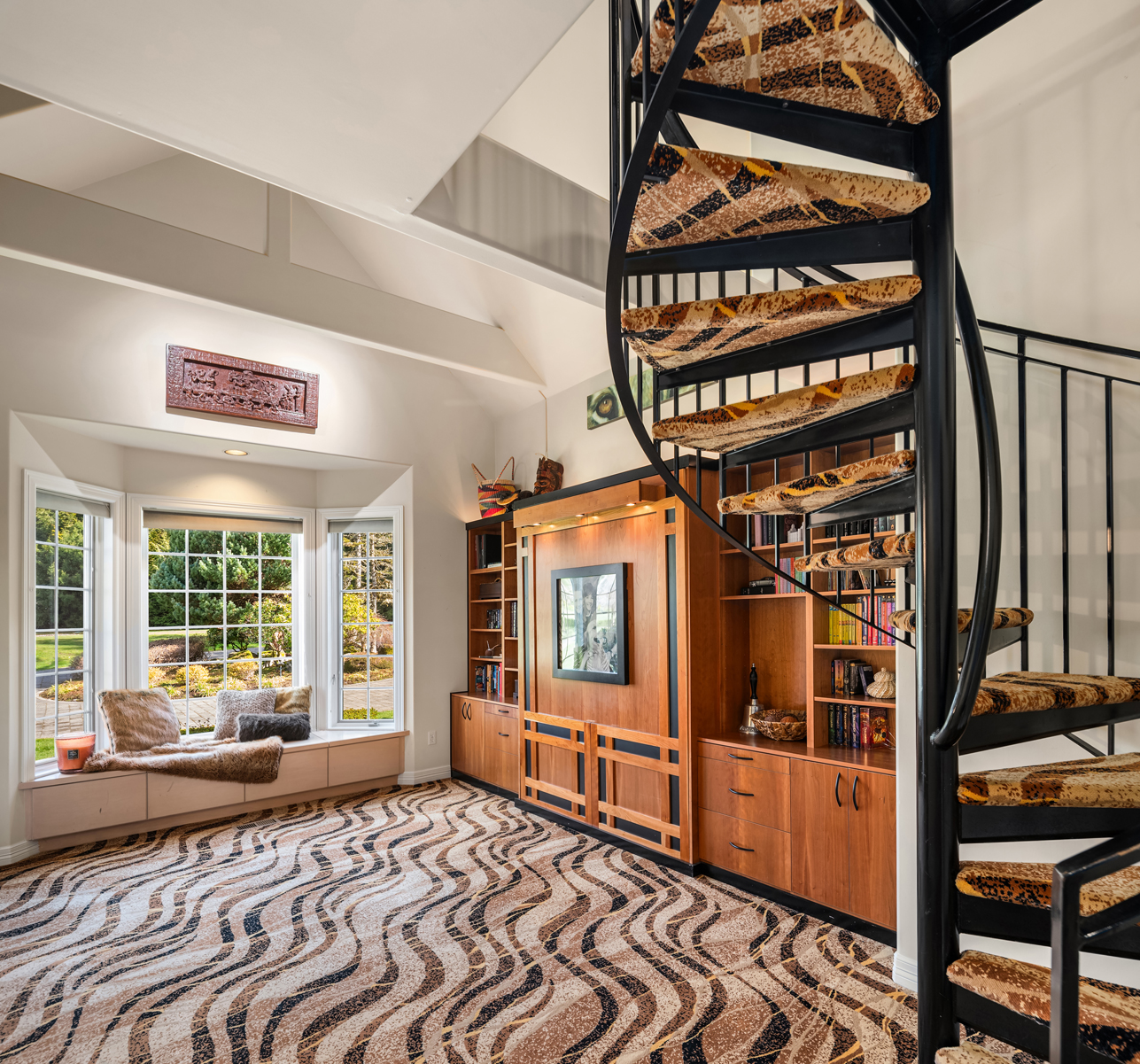 This living room features a unique spiral staircase, a built-in entertainment center with wooden cabinetry, and a cozy window seat with pillows. The room is well-lit with natural light from the bay window, and the patterned carpet adds visual interest. The overall impression is a blend of comfort and distinctive architectural design.