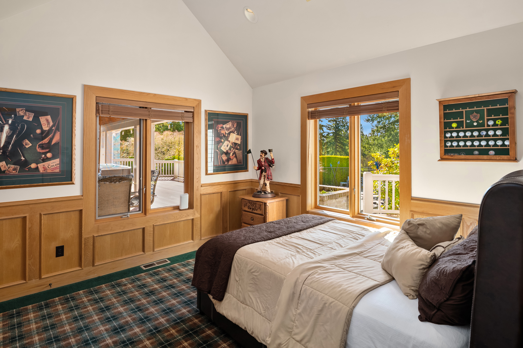 This is a cozy primary bedroom featuring wood-paneled walls and a plaid carpet. The room has two windows that let in natural light, with views of the outdoor greenery and a deck. The bed is neatly made with a brown and beige comforter and several pillows, creating a warm and inviting atmosphere.