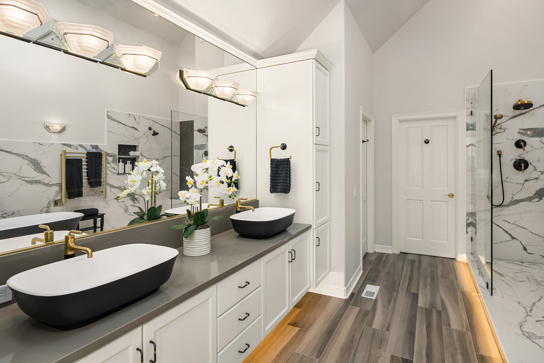 This is a primary bathroom featuring a double vanity with black and white vessel sinks, gold fixtures, and a large mirror with modern lighting. The bathroom has a clean, contemporary design with white cabinetry, gray countertops, and wood-look tile flooring. A glass-enclosed shower with marble-look tile is visible in the background, adding a luxurious touch.