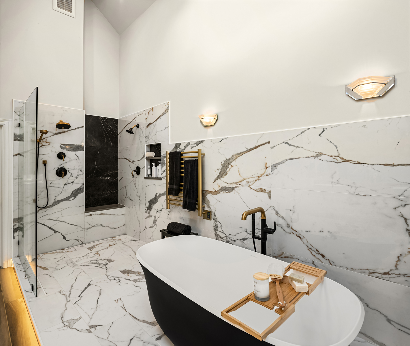 This is a luxurious primary bathroom featuring marble tiling on the floor and walls, creating a sophisticated and elegant ambiance. The bathroom includes a modern black and white freestanding bathtub with a wooden tray, a glass-enclosed shower with black fixtures, and a heated towel rack. The lighting is soft and warm, enhancing the spa-like atmosphere.
