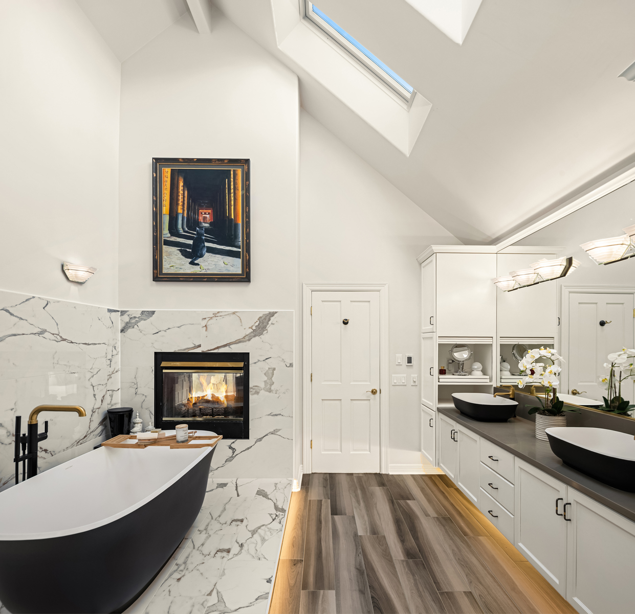 This is a luxurious primary bathroom featuring a freestanding black bathtub with a gold faucet, set against a marble-tiled wall with a fireplace. The room also includes a double vanity with black vessel sinks, white cabinetry, and modern lighting fixtures. The flooring is a wood-look plank, and a skylight provides natural light, creating a spa-like atmosphere.